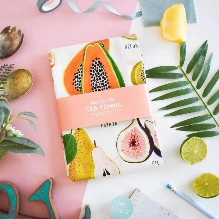Fruit design tea towel on pink background surrounded by fruit and leaves.