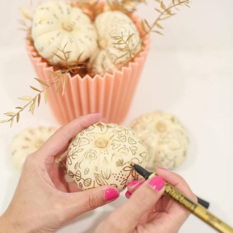 Hand holding small white pumpkin and drawing on pattern with gold pen.