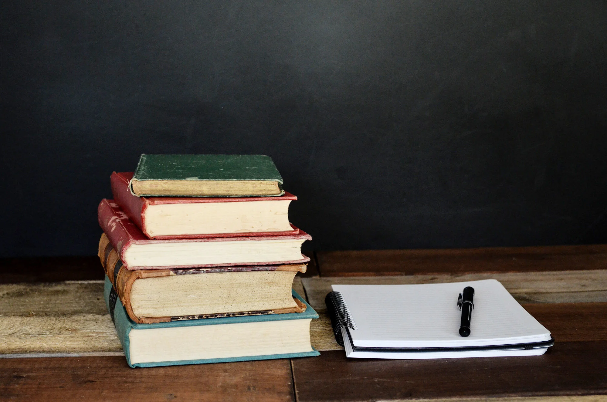 Old hardbacks sit next to a blank notepad and pen resting on a hardwood table.