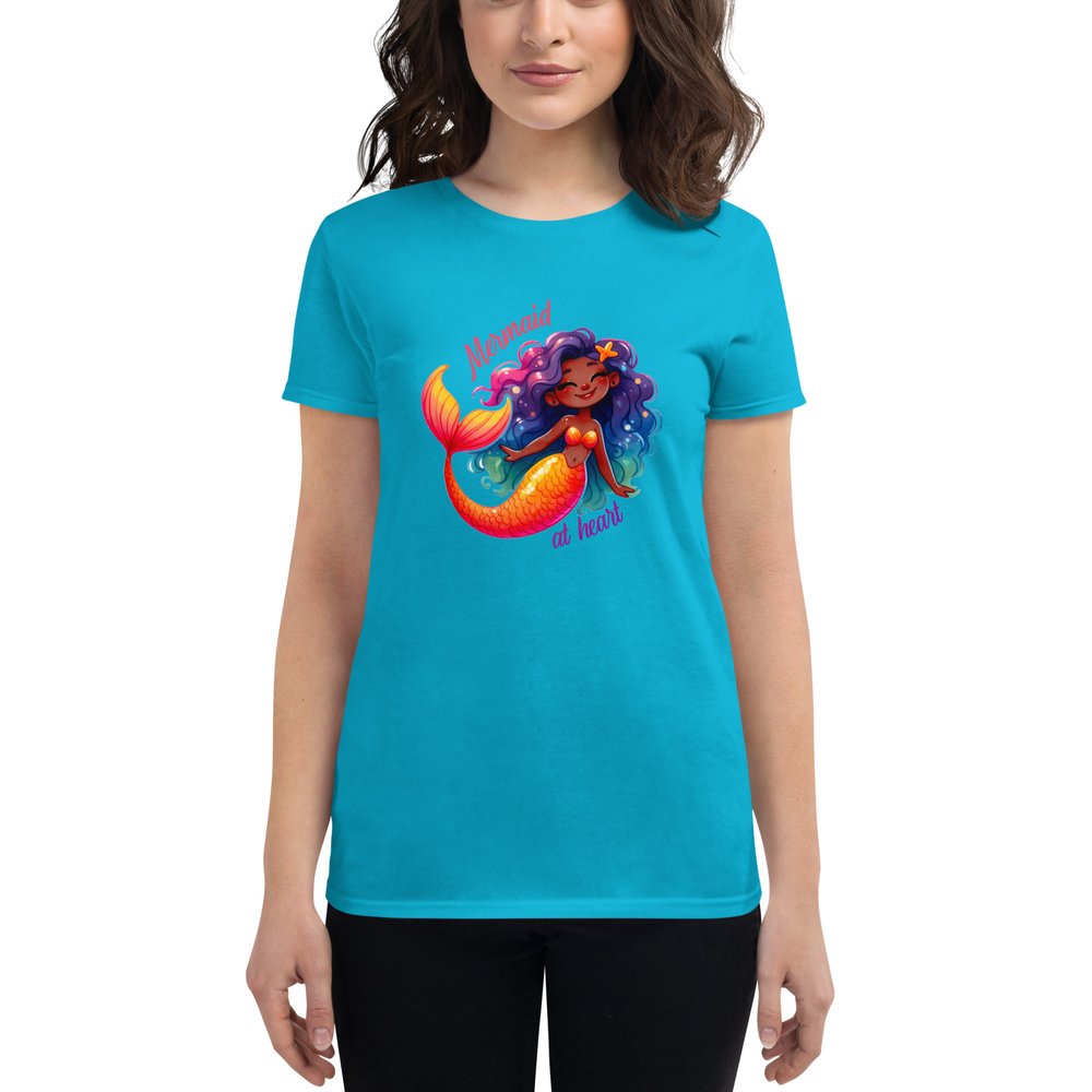 A woman in dark hair wears a Caribbean blue short-sleeved T-shirt with a large colourful graphic showing a sweet Black mermaid with an orange tail and long multi-coloured hair. Text: Mermaid at heart.
