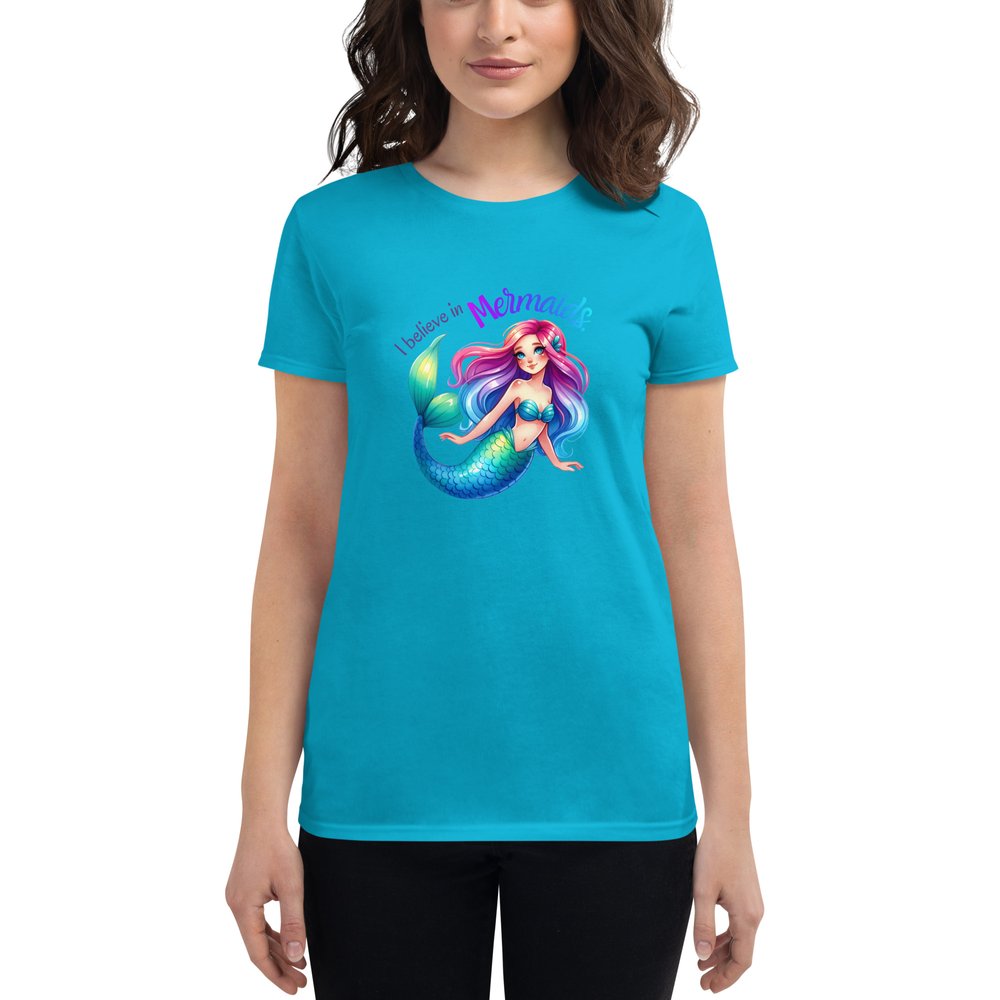 Woman wearing a Caribbean blue short-sleeved T-shirt with a large centered graphic of a Caucasian mermaid with rainbow hair, clam shell bra, and a blue-green tail. Text: I believe in mermaids.