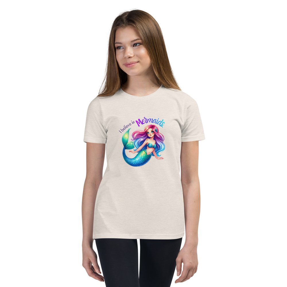 White pre-teen girl wears a heather dust colour youth tee with a colourful mermaid graphic and text that reads "I believe in mermaids."