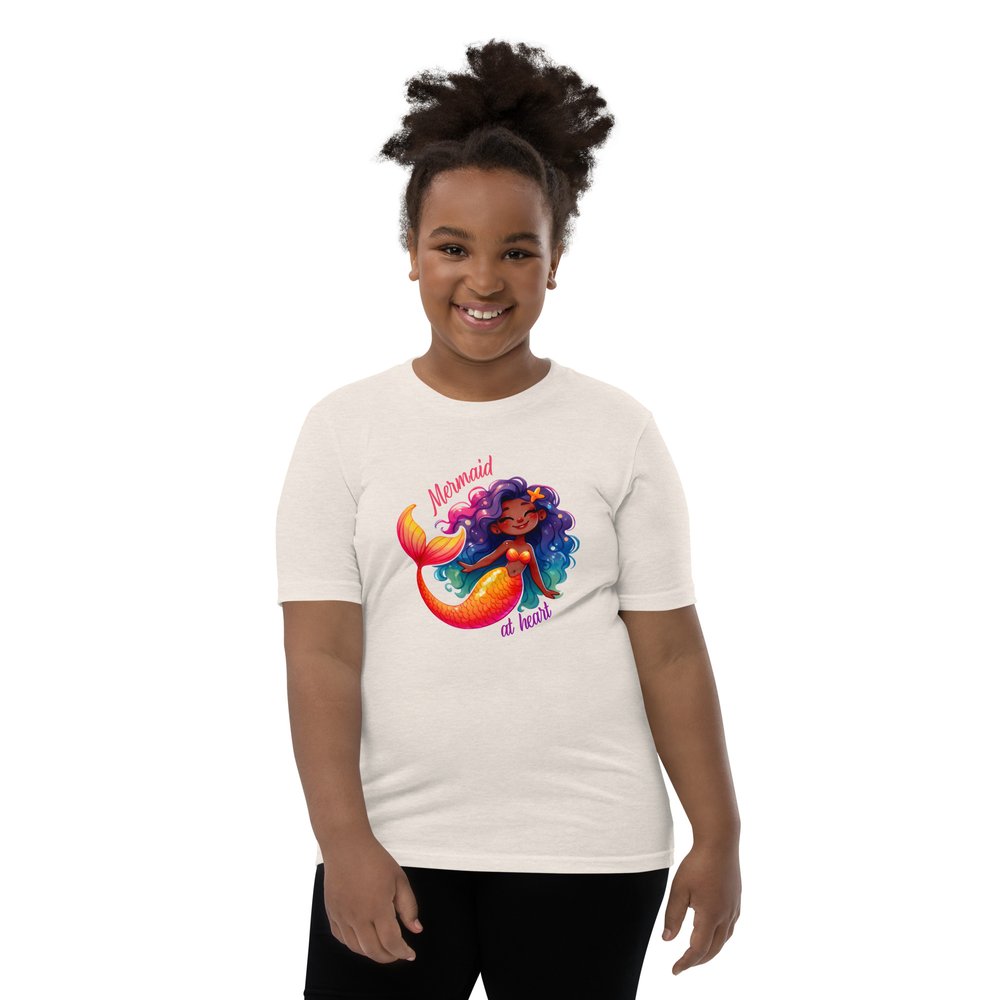 Pre-teen Black girl wearing a heather dust youth tee with a colourful, cute Black mermaid girl graphic and text "Mermaid at heart".