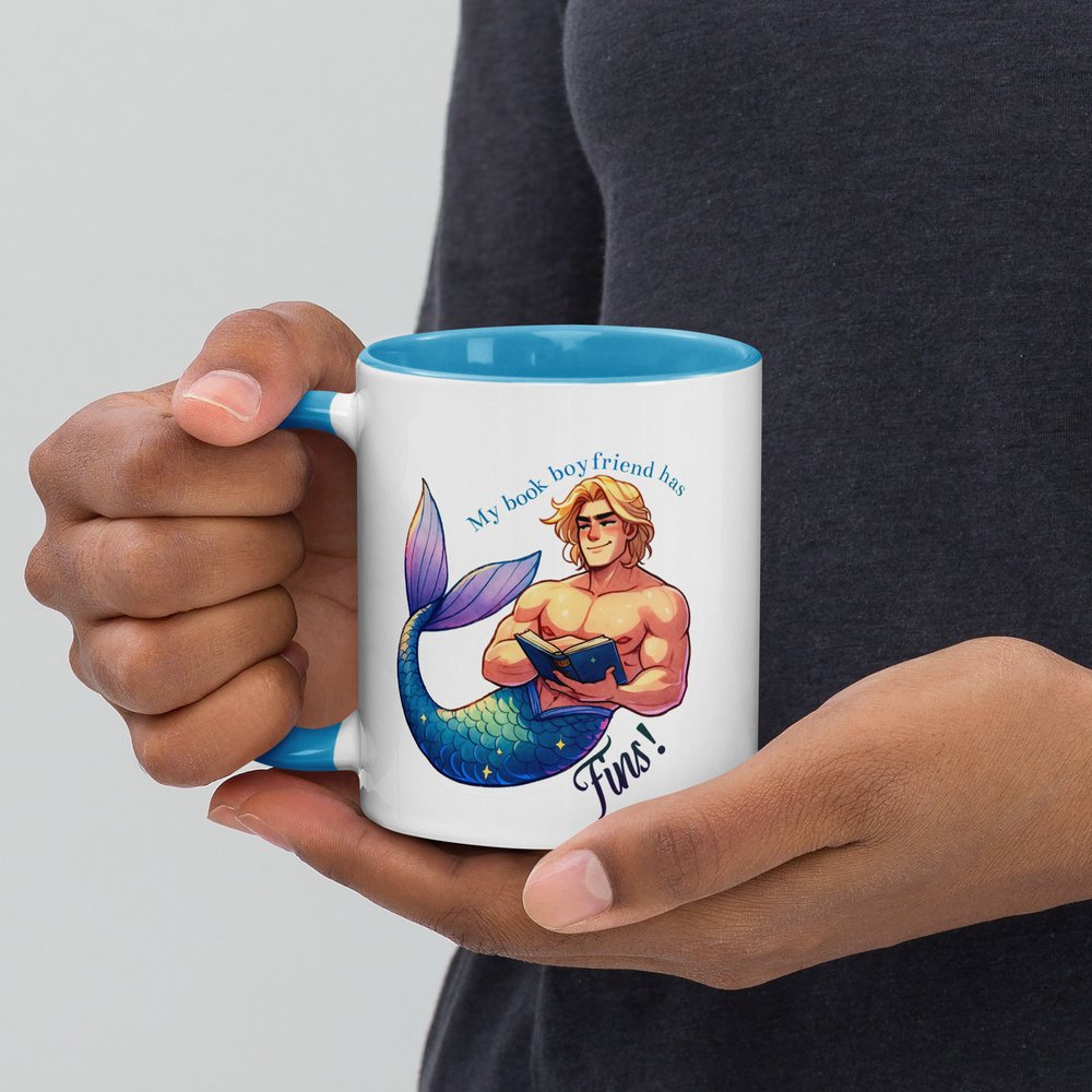 A woman's hands hold an 11-ounce white ceramic mug with a bright blue interior and handle that has a colourful graphic shows a hunky blond merman reading and the text "My book boyfriend has fins!"