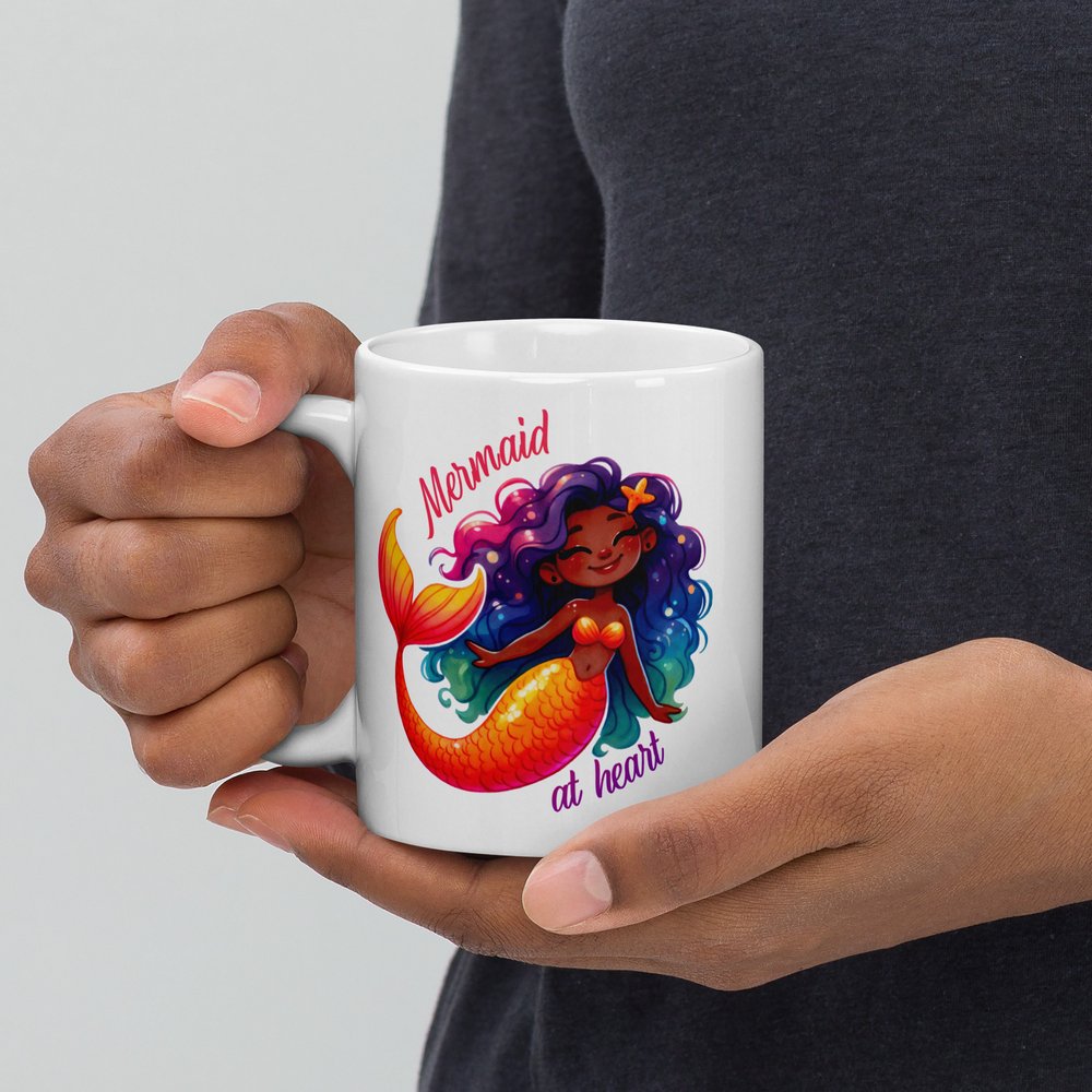 11-ounce white ceramic mug with handle on left showing a colourful Black mermaid graphic with text that reads “Mermaid at heart”.