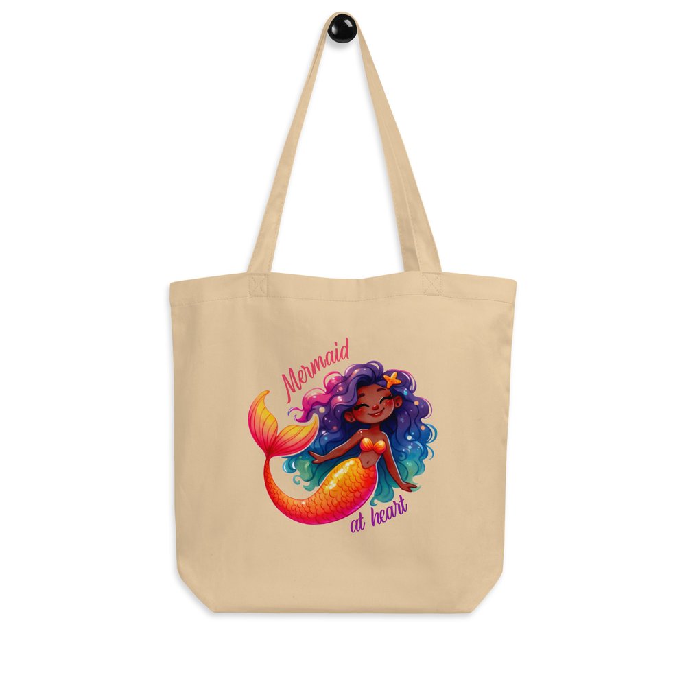 Front of a natural unbleached cotton tote hanging from a knob and showing a colourful graphic of a sweet and happy Black mermaid with the text "Mermaid at heart."