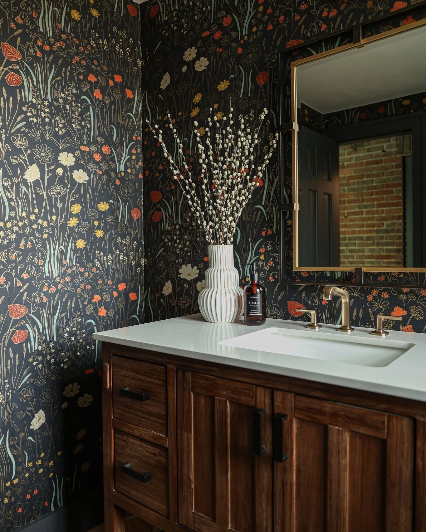 Moody powder room design with dark floral wallpaper, a dark stained wood vanity, brass faucet and brass framed mirror by a Tulsa interior designer