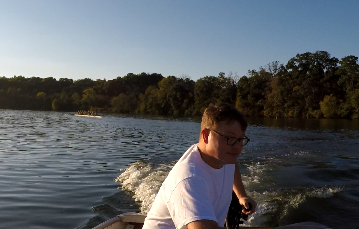 A person wearing glasses and a white t-shirt sitting in a boat on a river, with trees lining the riverbank and another boat in the distance.