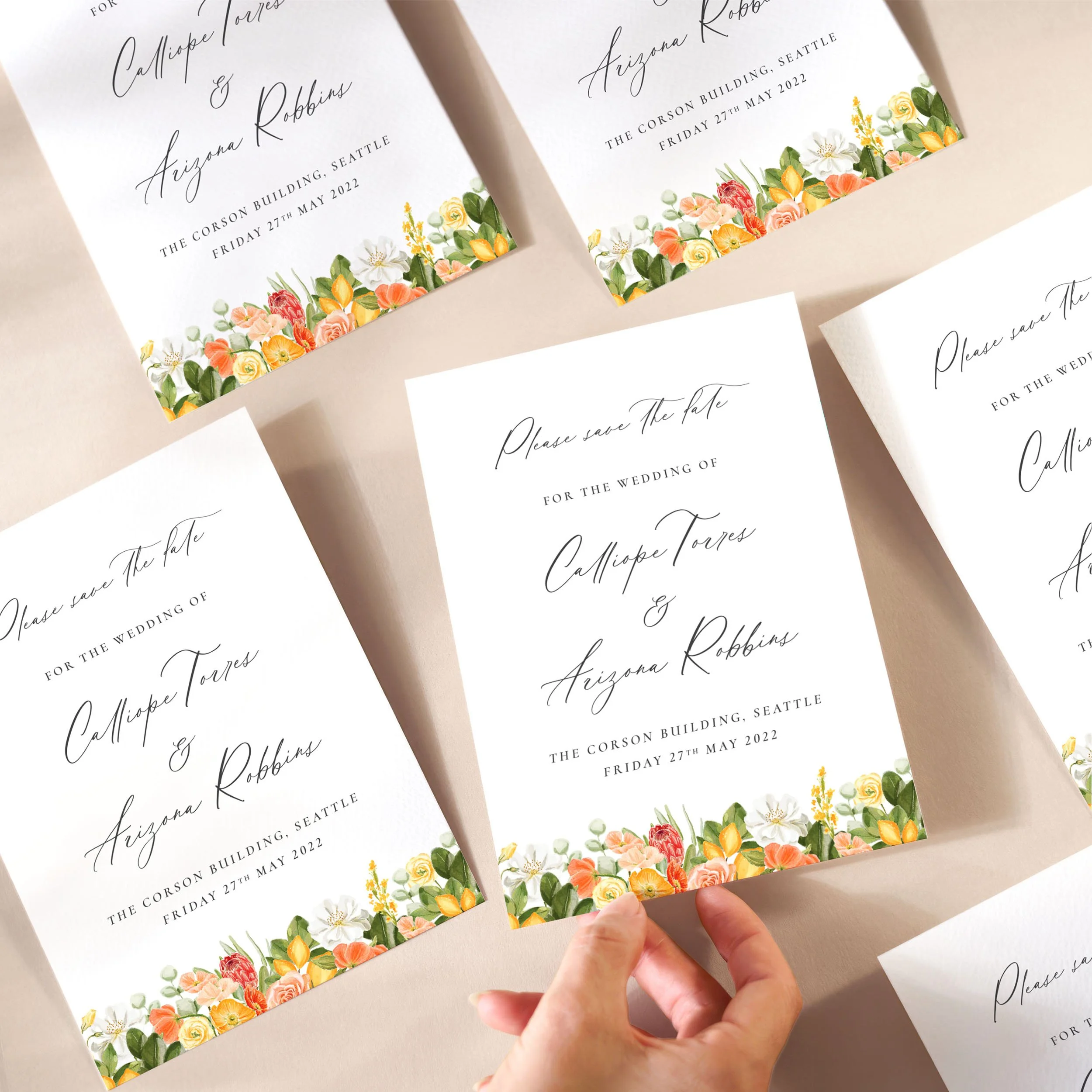 A hand picking up a save the date card with yellow and orange flowers, lemons along the bottom and a calligraphy style text.