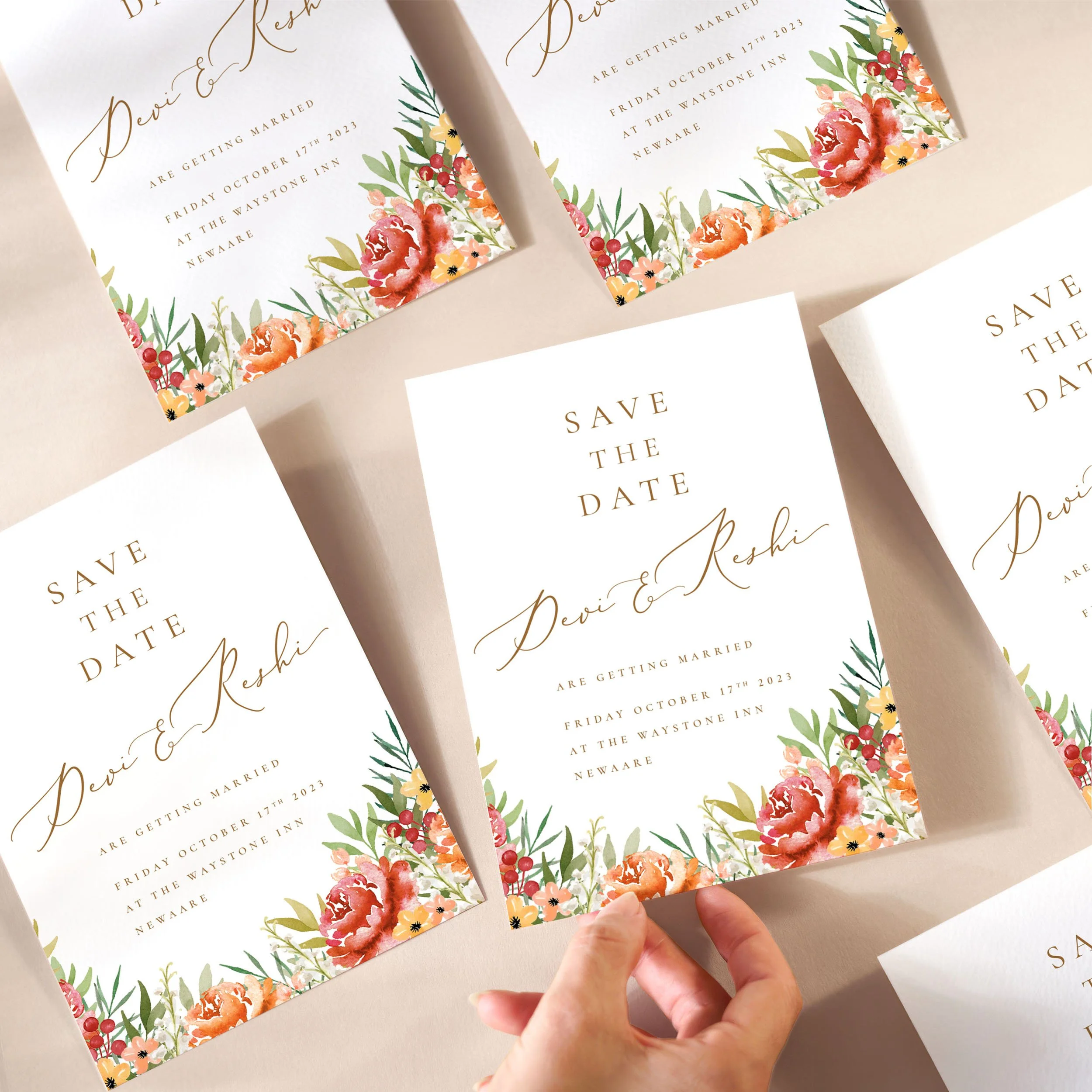 A hand picking up a save the date card with burgundy and orange flowers and gold calligraphy text.