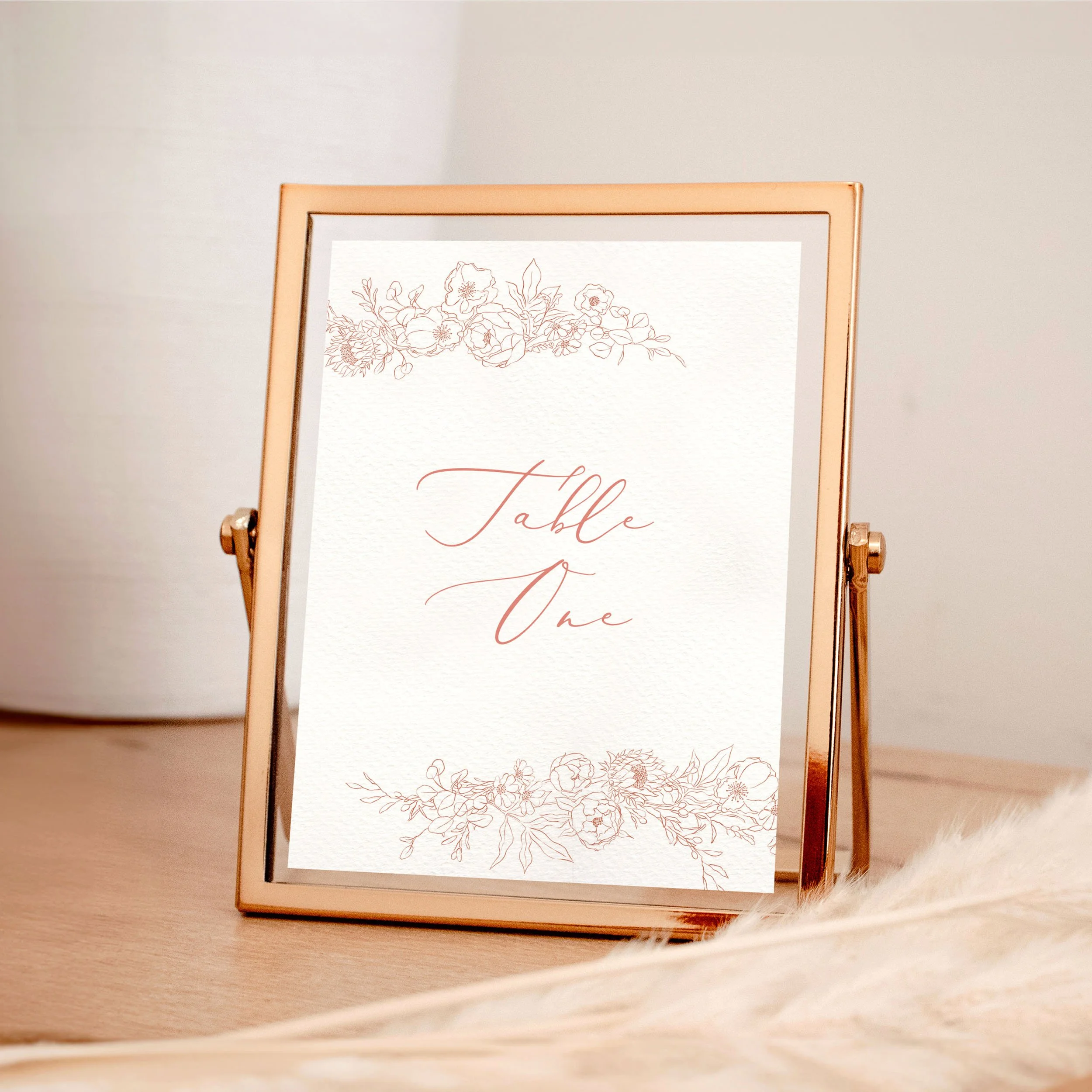 A table wedding number card in a rose gold frame with pink hand-drawn floral illustrations framing the edges