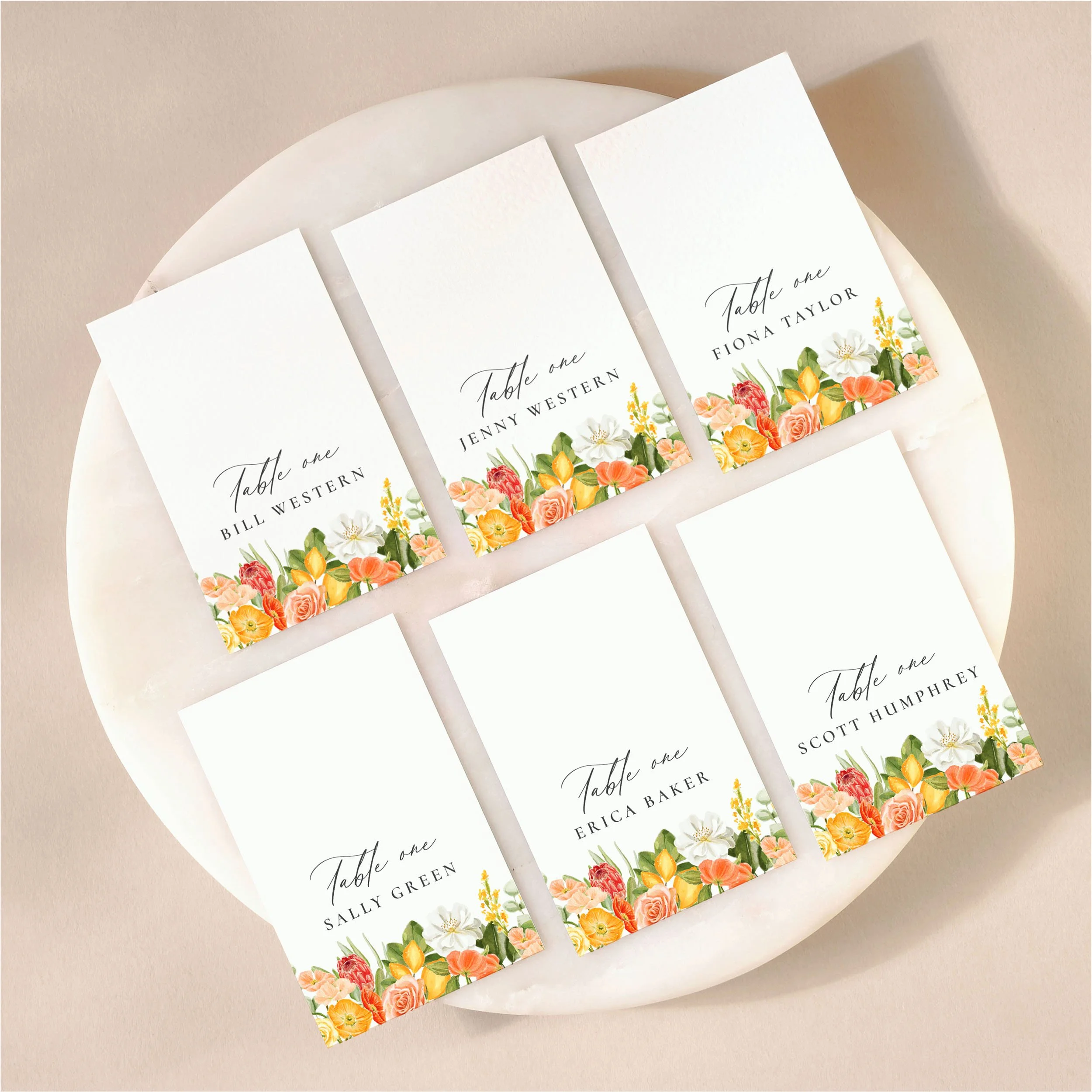 A set of 6 small, flat wedding place cards on a marble tile with printed guest names featuring illustrations of yellow and orange flowers as well as some small lemons.