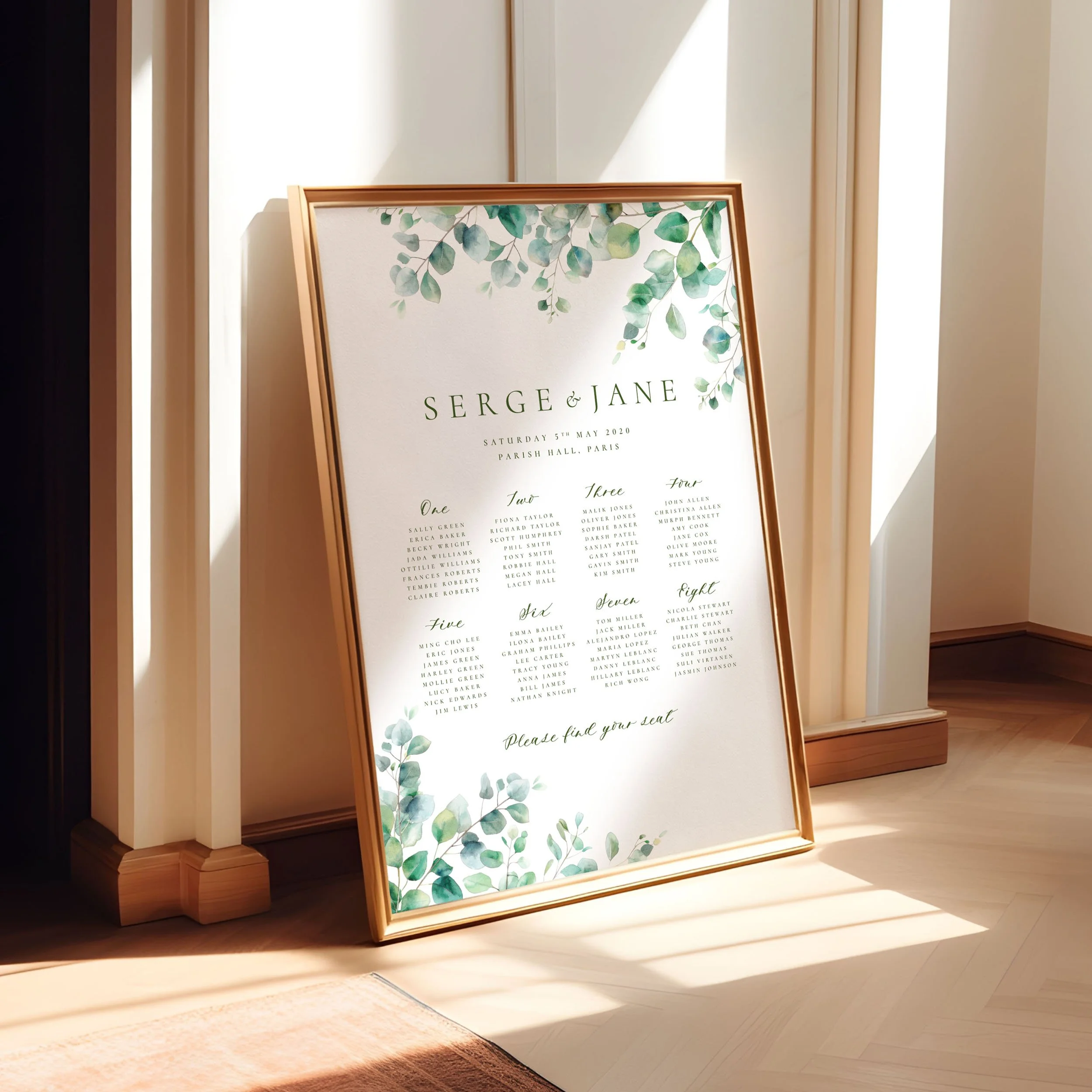 A framed A1 table plan sign leaning against a wall as the light spills across. The design has watercolour eucalyptus leaves spilling from two corners.
