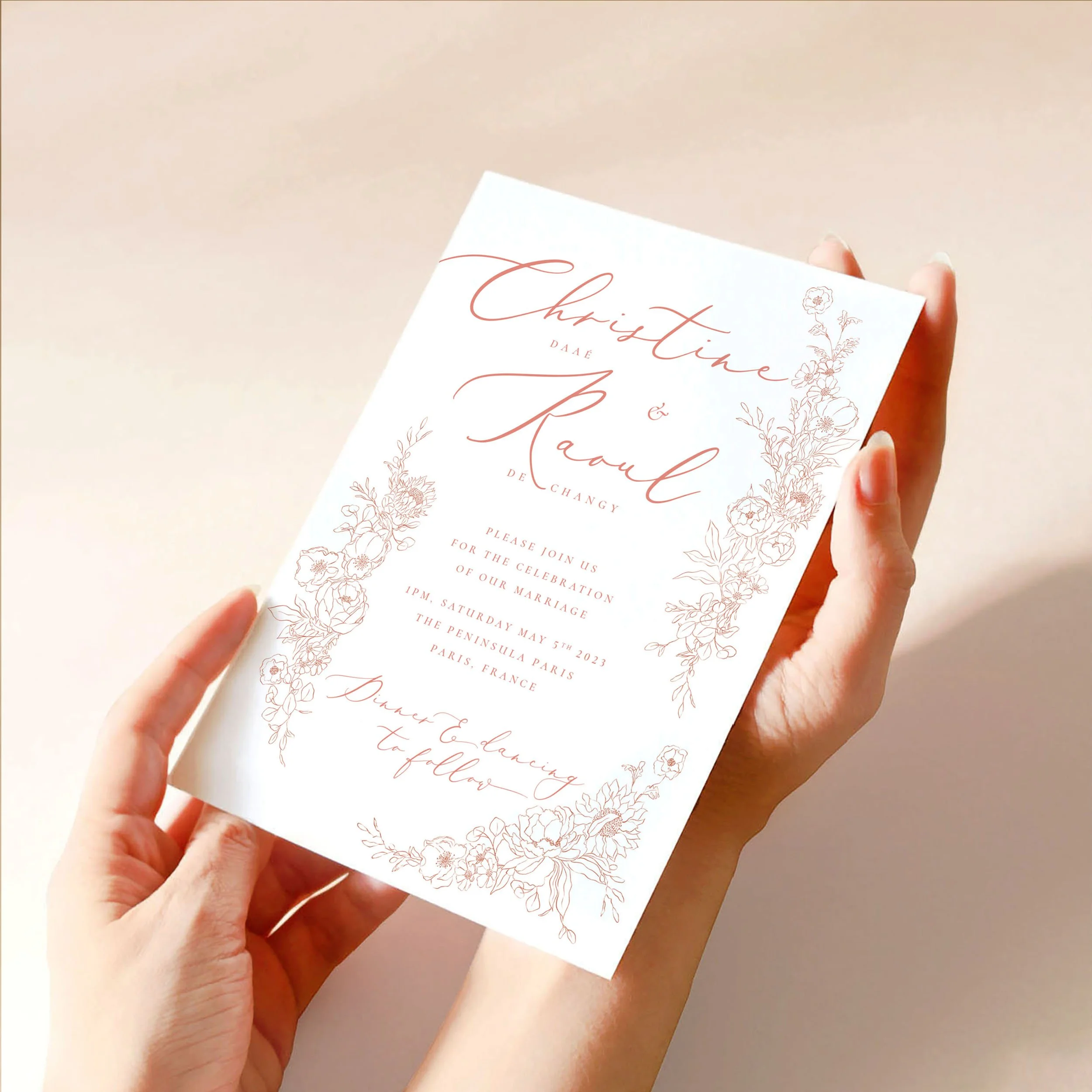 Two hands hold a 5x7" wedding invitation card. The card has a soft pink hand-drawn floral design framing blush pink, calligraphy-style text.