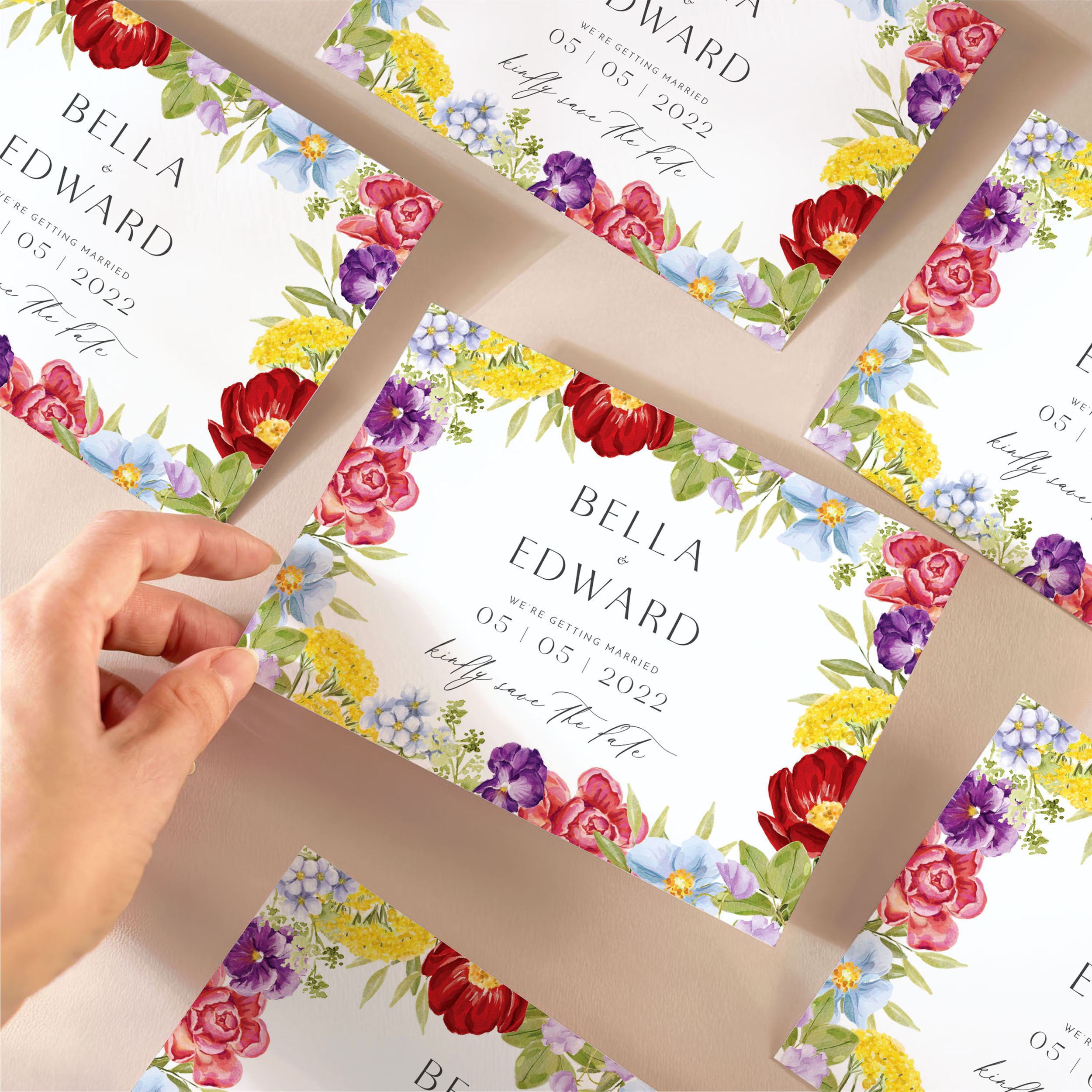 A hand picking up a save the date card with bright watercolour flowers around the edges, and a modern mix of serif and calligraphy-style fonts.