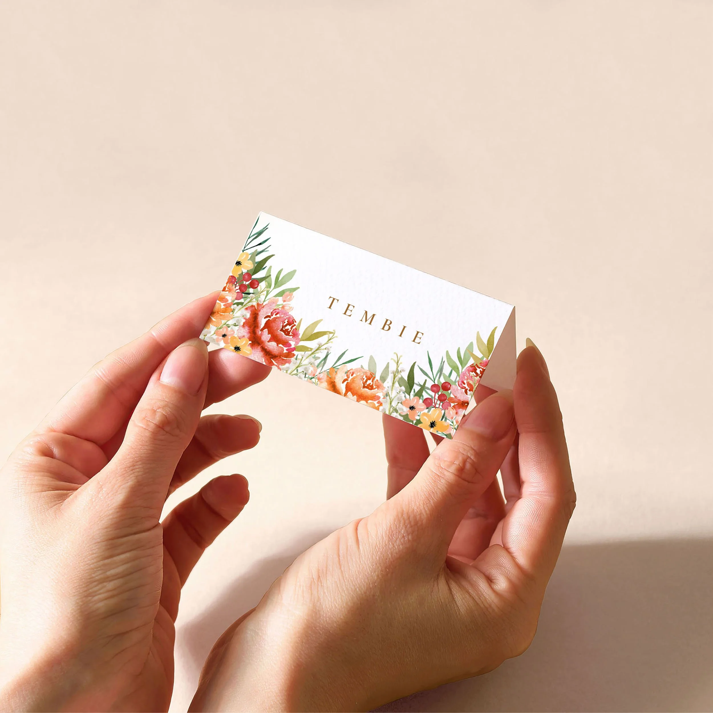 Hands holding a white tent fold place card with a guest name printed on it and warm watercolour flowers in shades of red and orange.