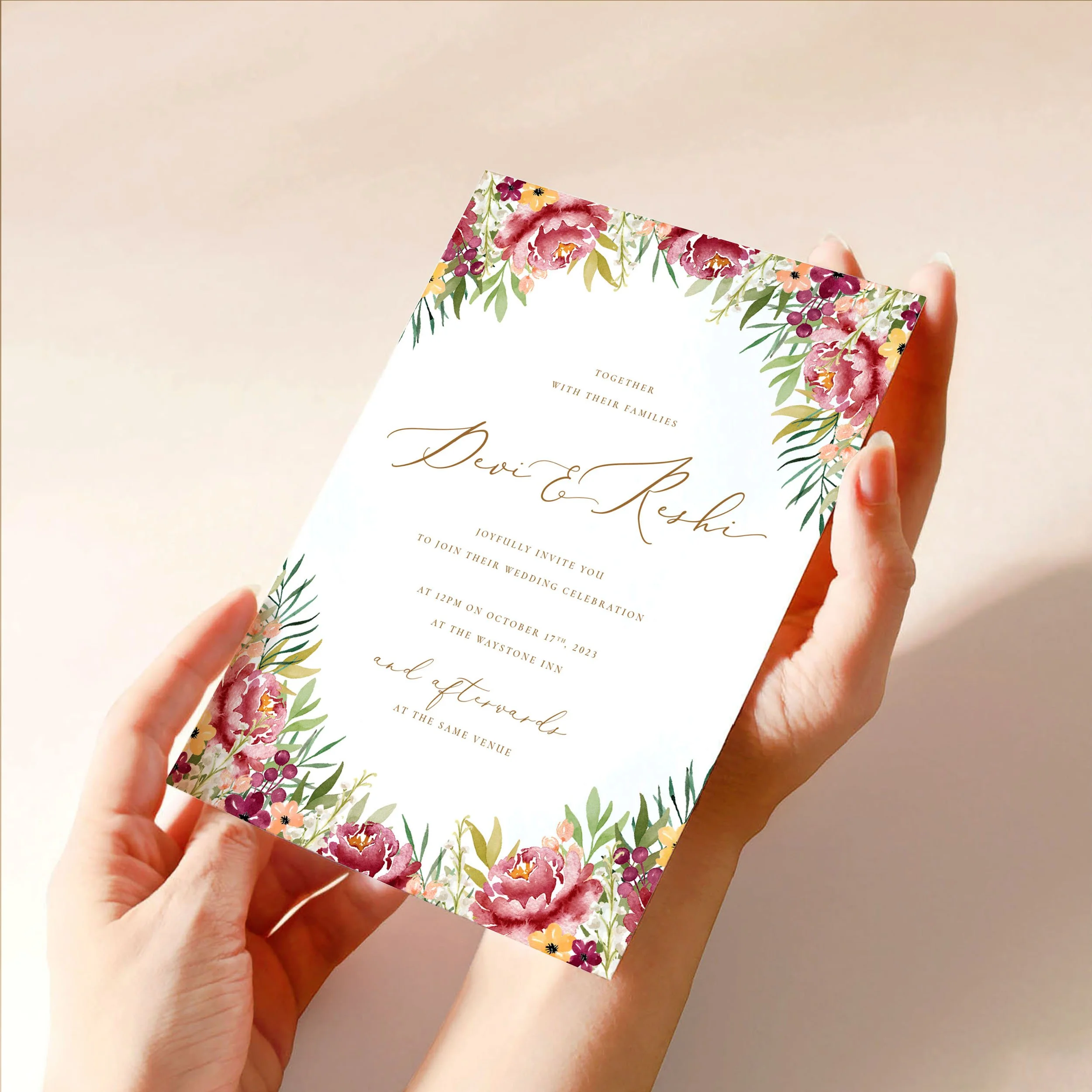 Two hands hold a 5x7" wedding invitation card. The card has a soft burgundy watercolour floral design with green leaves around the edges and gold text.