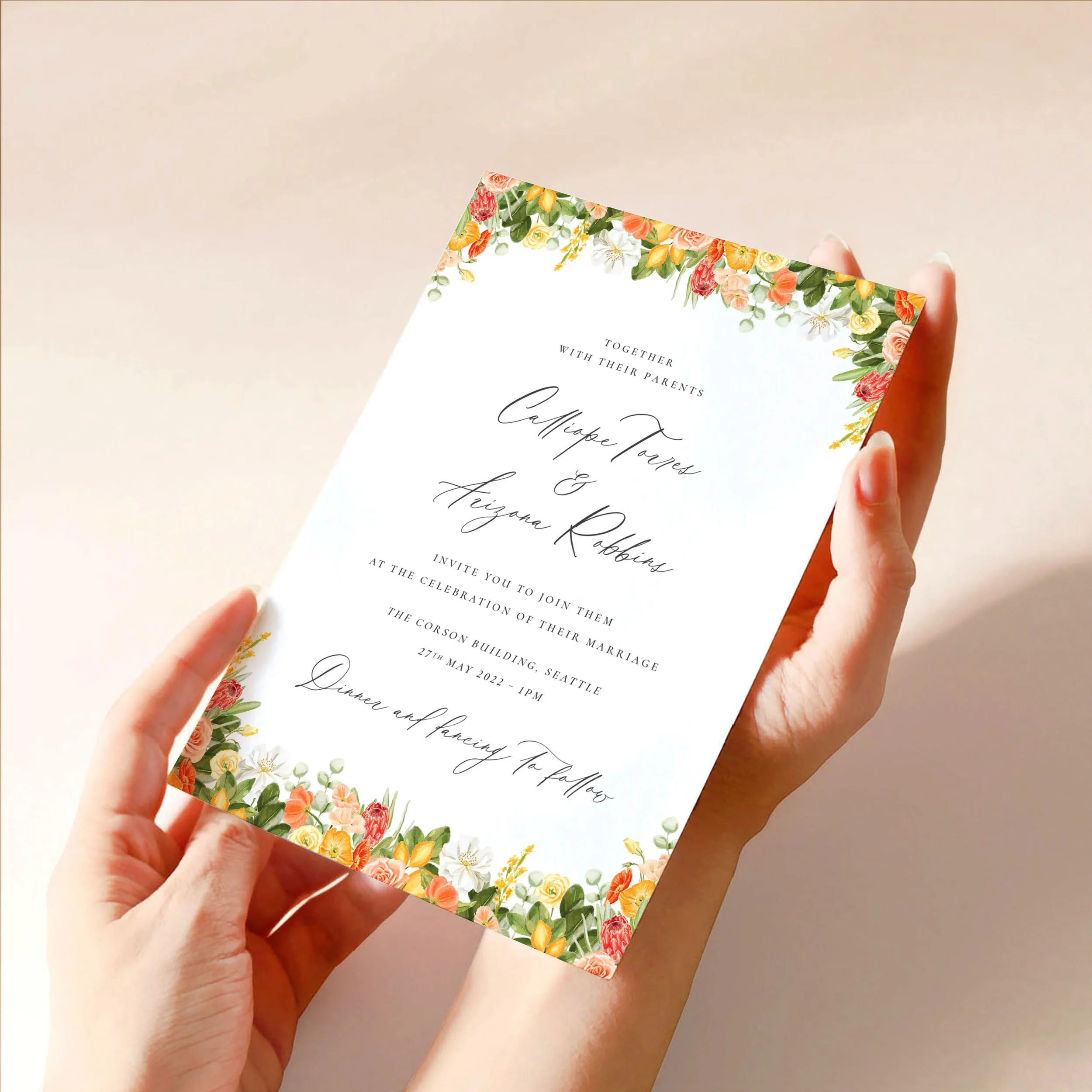 Two hands hold a 5x7" wedding invitation card. The card has a hand-painted yellow and orange citrus design at the top and bottom and calligraphy-style text.