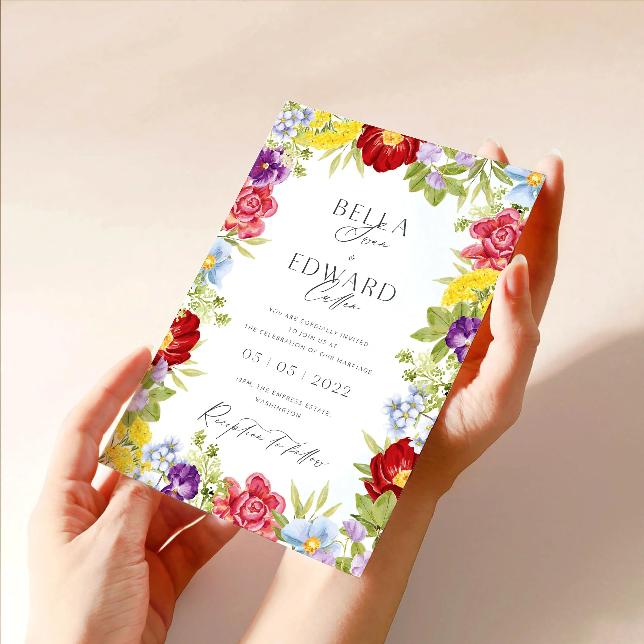 Two hands hold a 5x7" wedding invitation card. It has a selection of bright hand-painted flowers around the edges, framing modern and calligraphy-style text.
