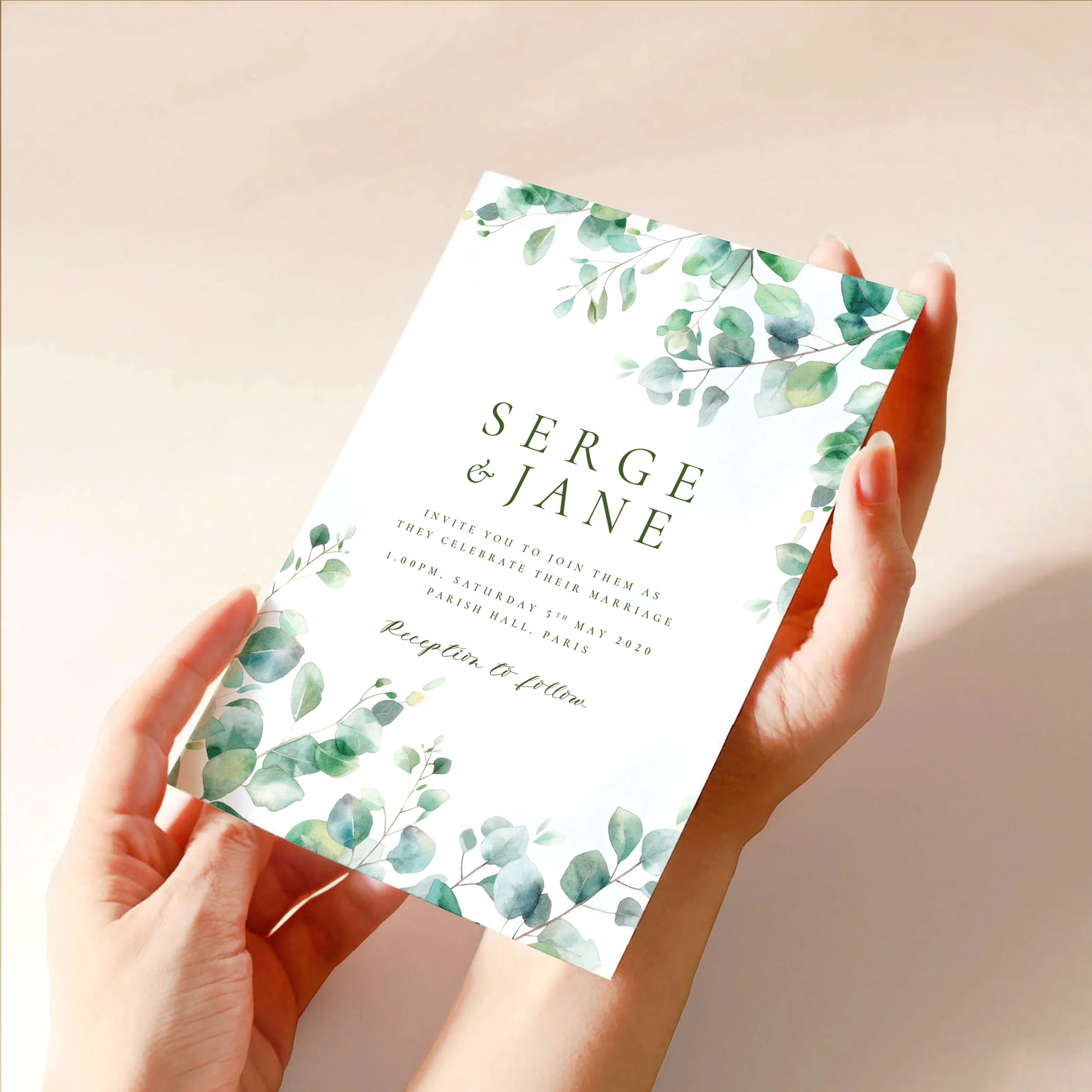 Two hands hold a 5x7" wedding invitation card. The design has soft green eucalyptus leaves spilling in from two corners and modern font choice.