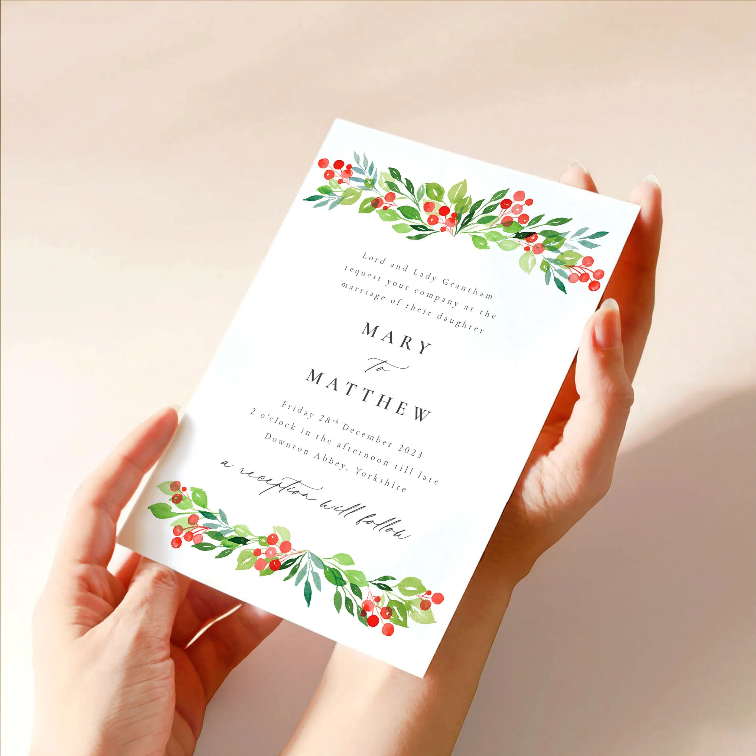 Two hands hold a 5x7" wedding invitation card. It has a Christmas wreath style hand-painted design at the top and bottom with red berries and green leaves.