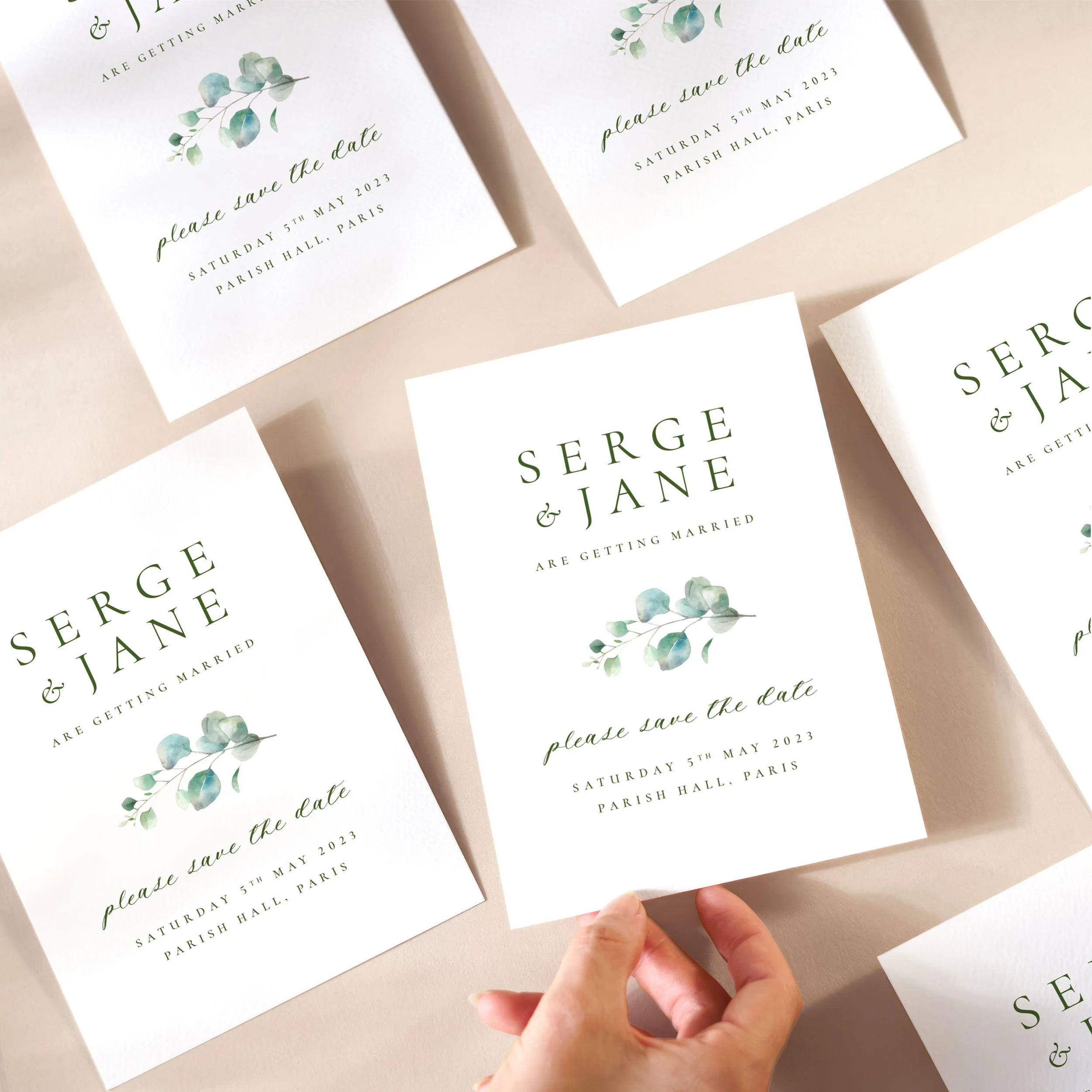 A hand picking up a save the date card with a watercolour eucalyptus leaf, and a modern mix of serif and handwritten-style fonts.