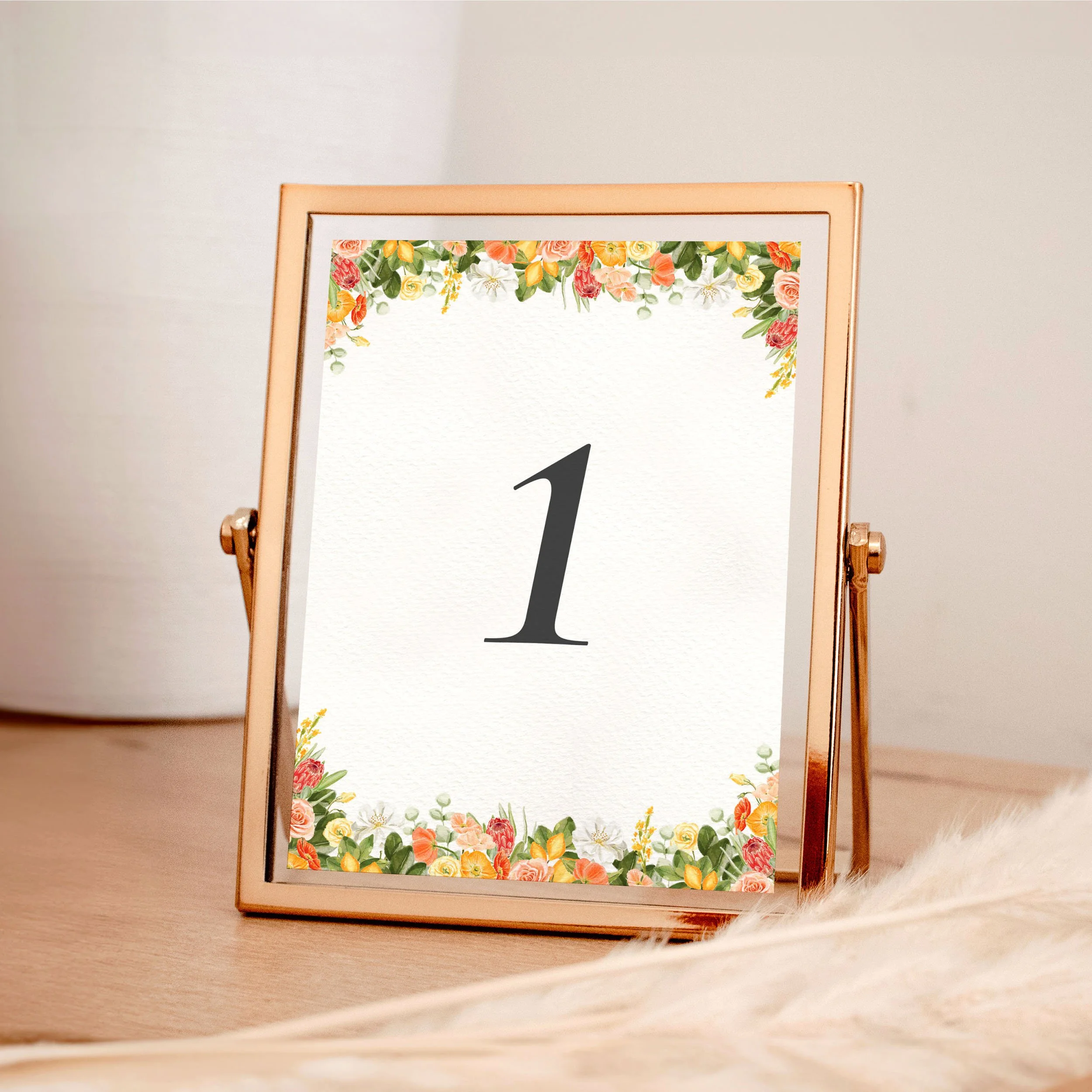 A table wedding number card in a rose gold frame with yellow and orange flowers framing the top and bottom, along with green leaves.