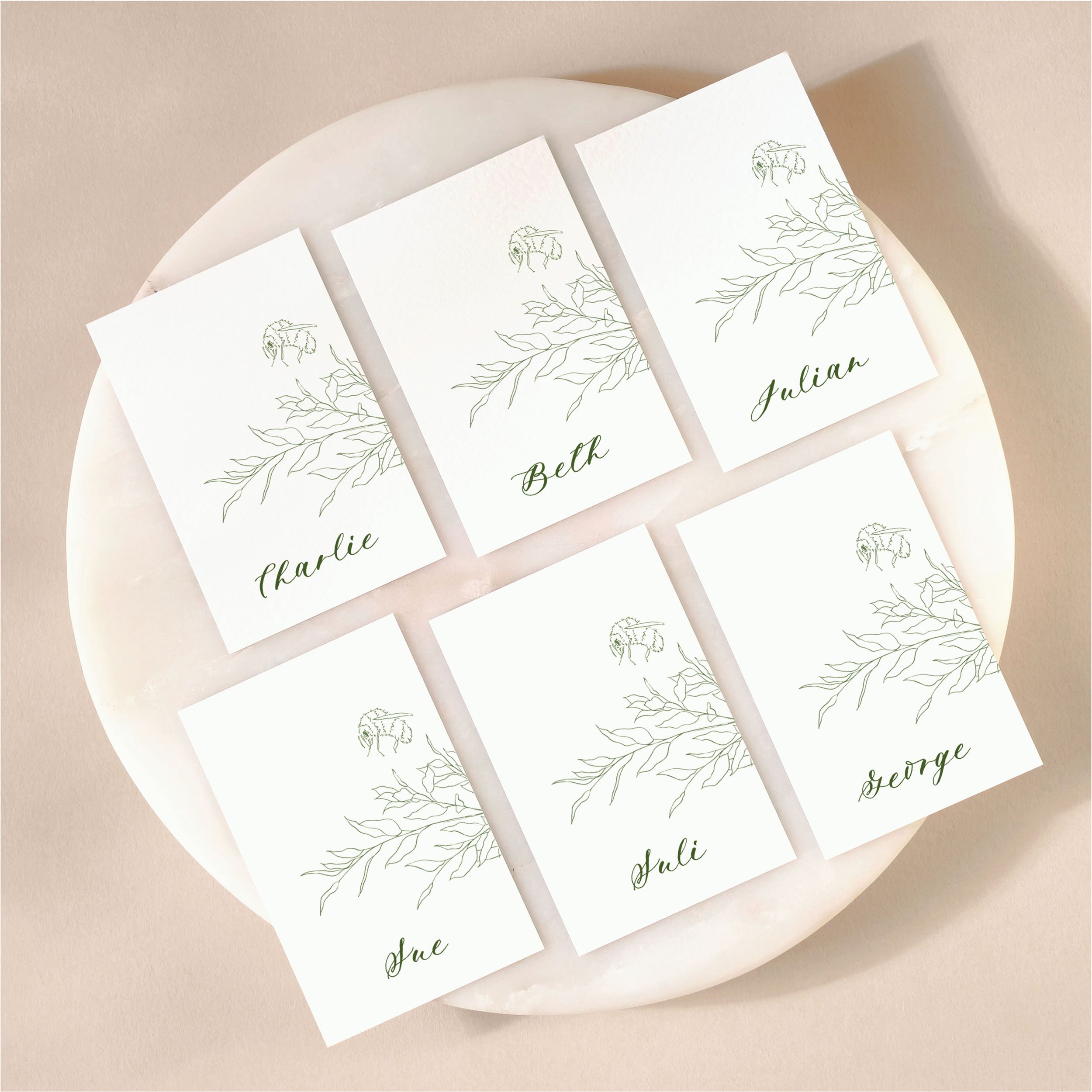 A set of 6 small, flat wedding place cards on a marble tile with printed guest names featuring an illustration of some leaves and a bumble bee