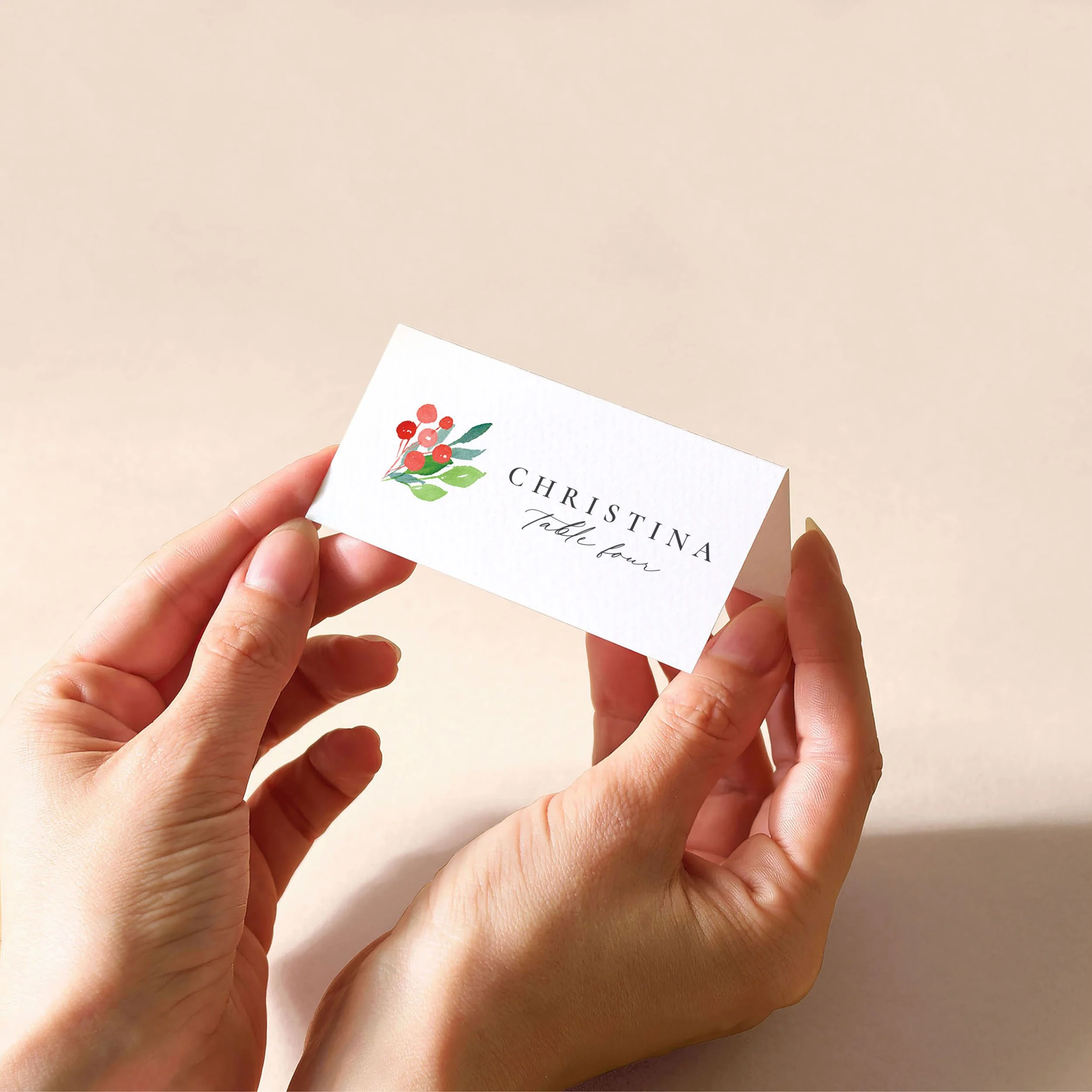 Hands holding a white tent fold place card with a guest name printed on it and a design of small watercolour winter berries.