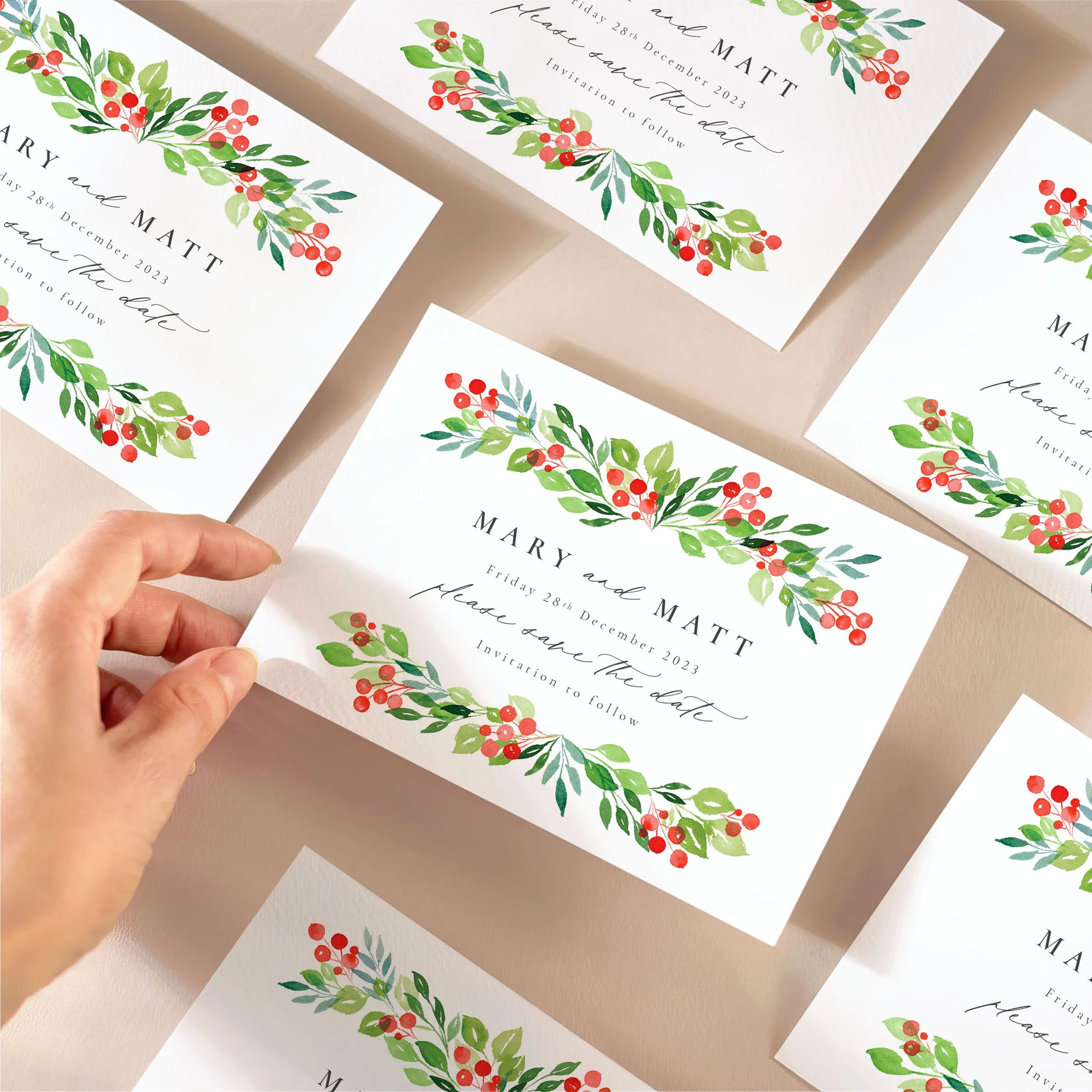 A hand picking up a save the date card with watercolour berries and green leaves at the top and bottom, and a mix of serif and calligraphy-style fonts.