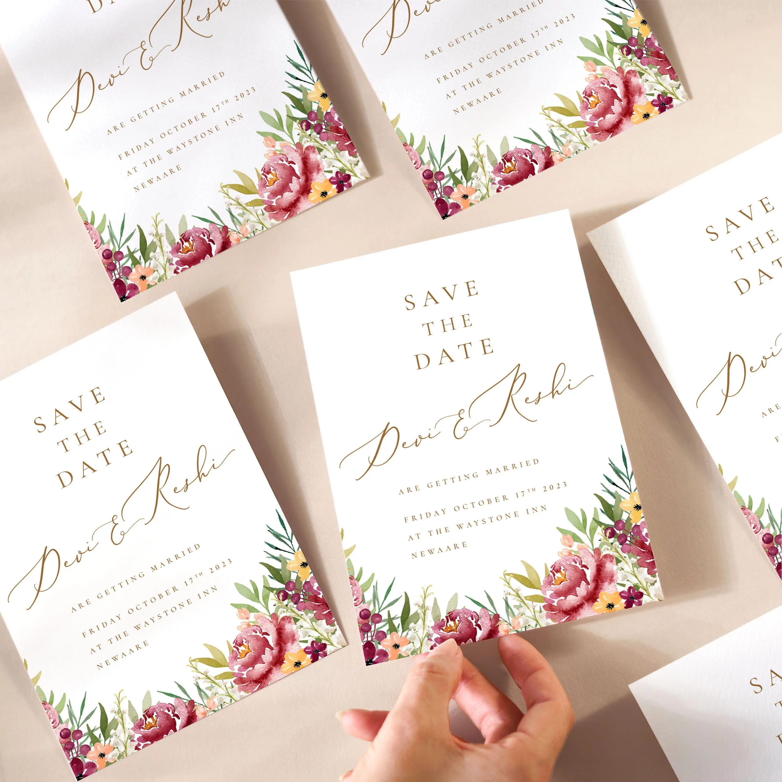 A hand picking up a save the date card with burgundy flowers and gold calligraphy text.