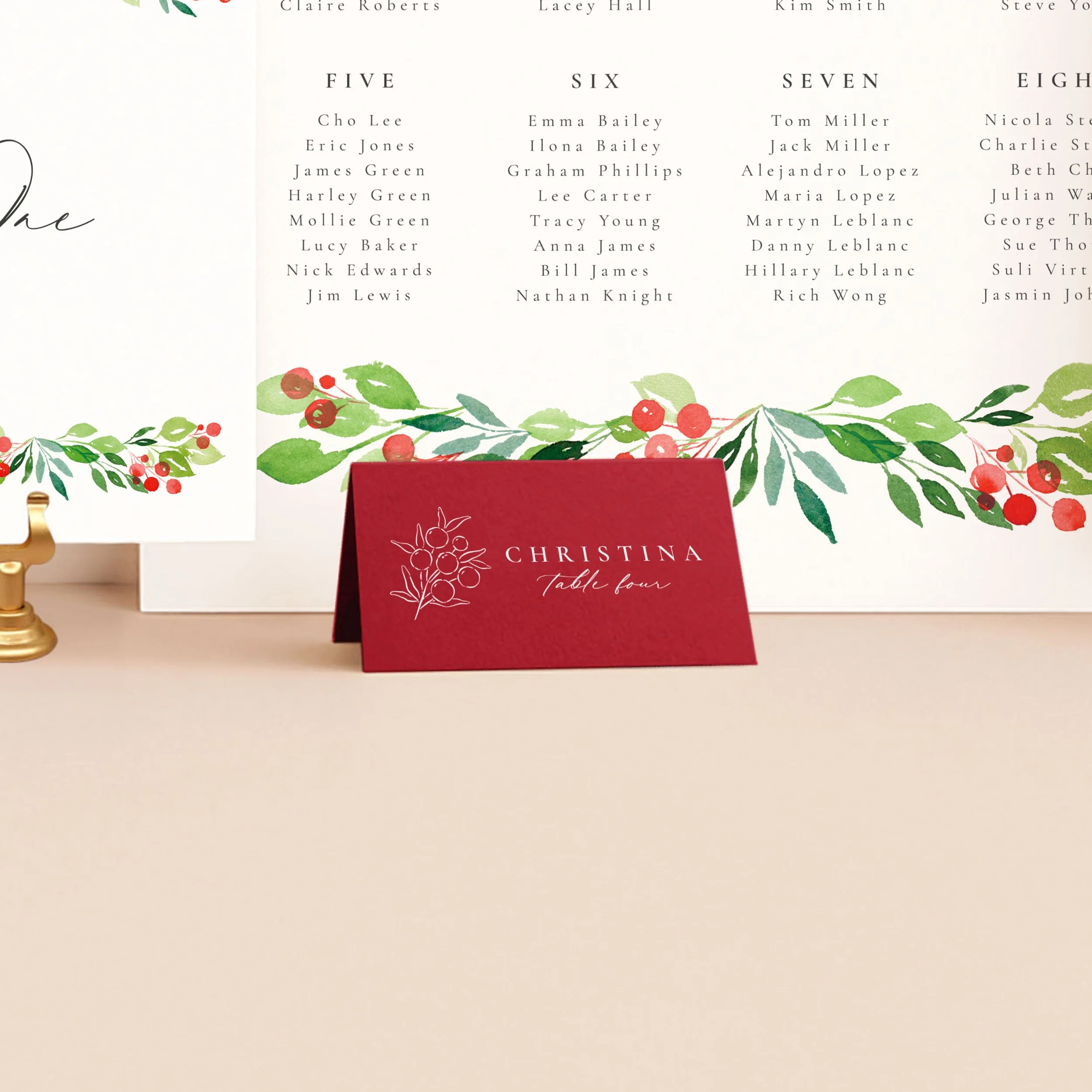 A cherry red tent place card, printed in white ink with a guest name and a winter berry sprig illustration.