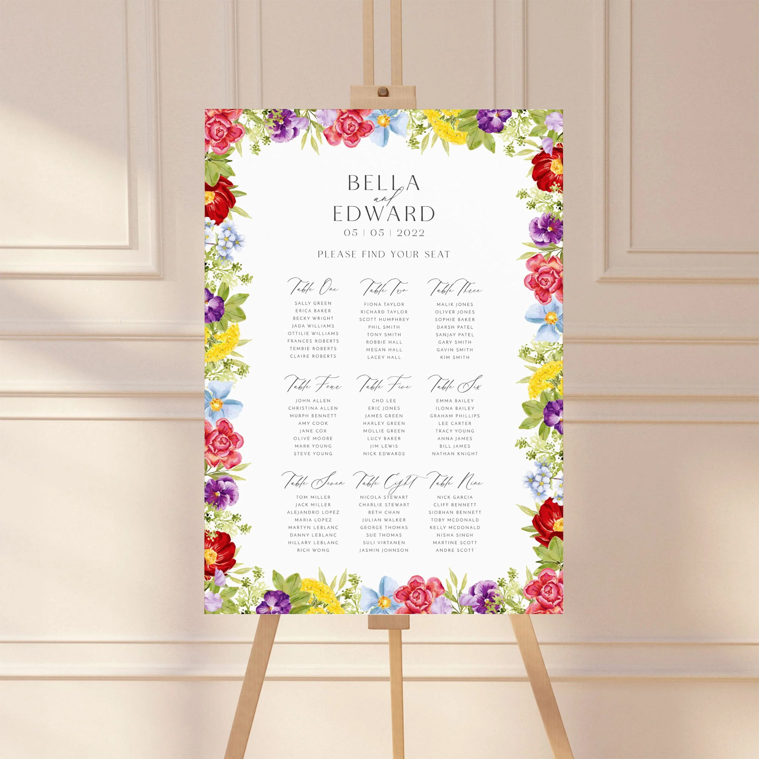 A wedding table plan sign printed on a large A1 board, standing on an easel. The design has bright watercolour florals in shades of red, pink, purple, blue, yellow and green.