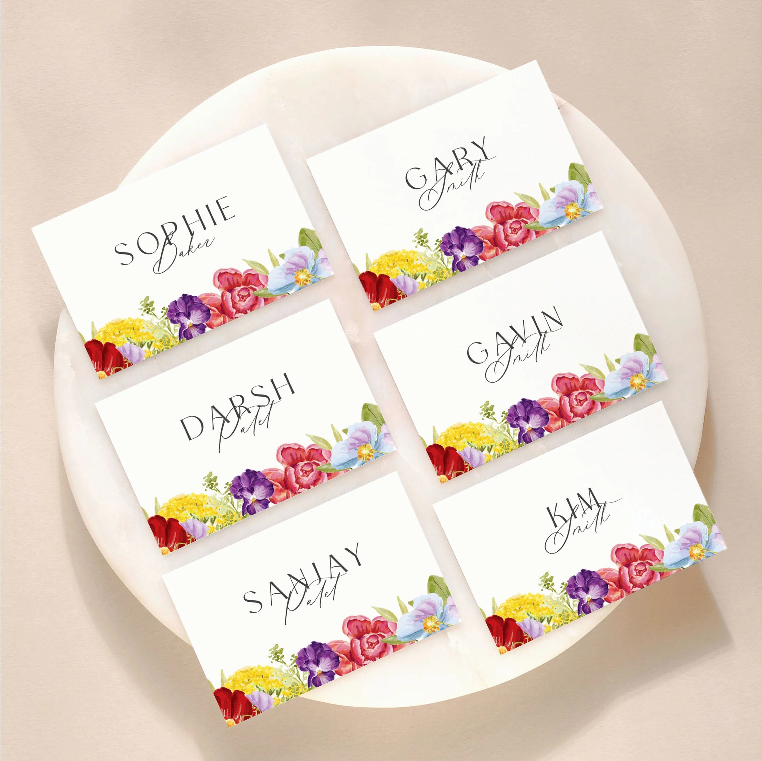 A set of 6 small, flat wedding place cards on a marble tile with printed guest names and illustrations of bright colourful flowers.