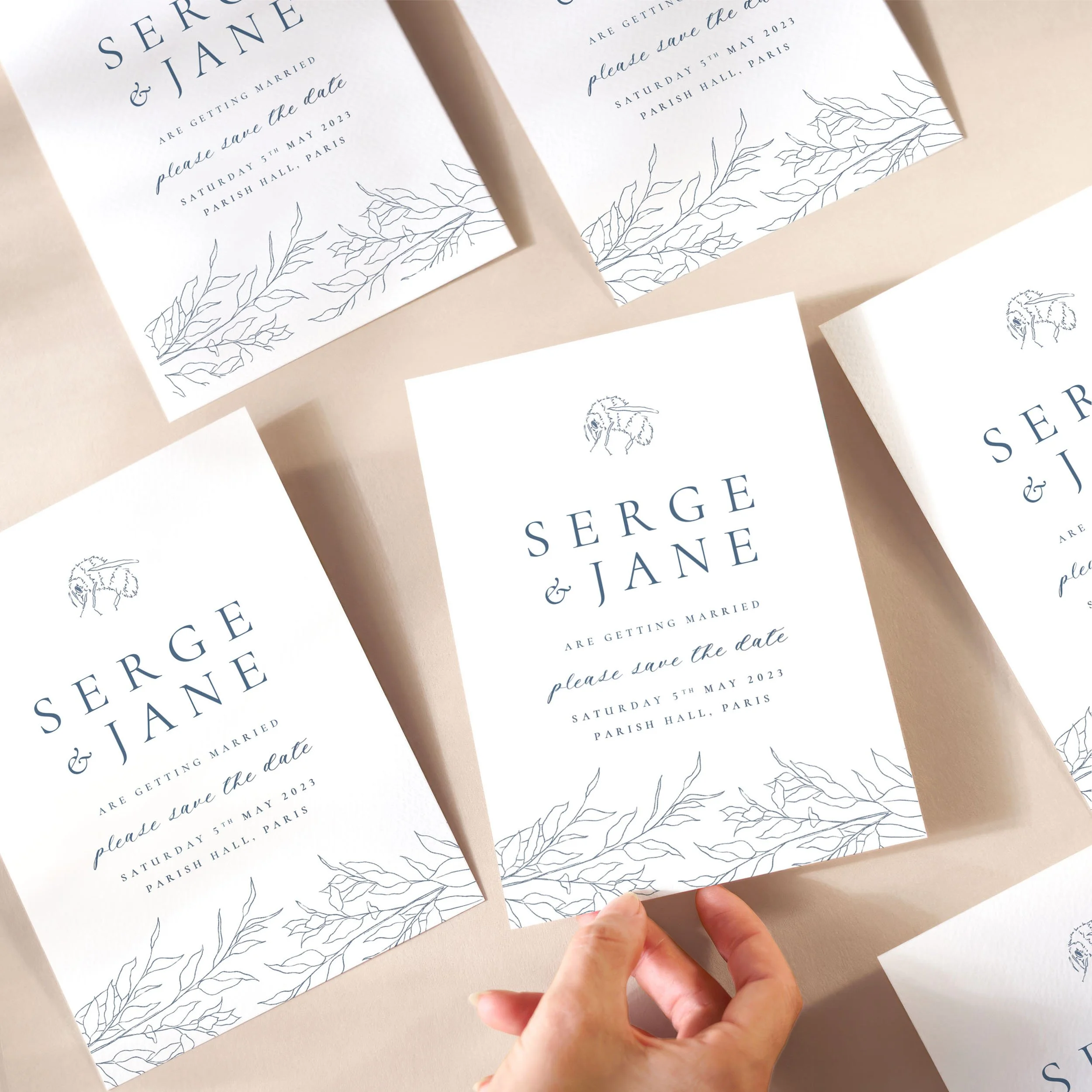 A hand picking up a save the date card with a delicate blue sketch and dusty blue serif text and a bumble bee illustration at the top.