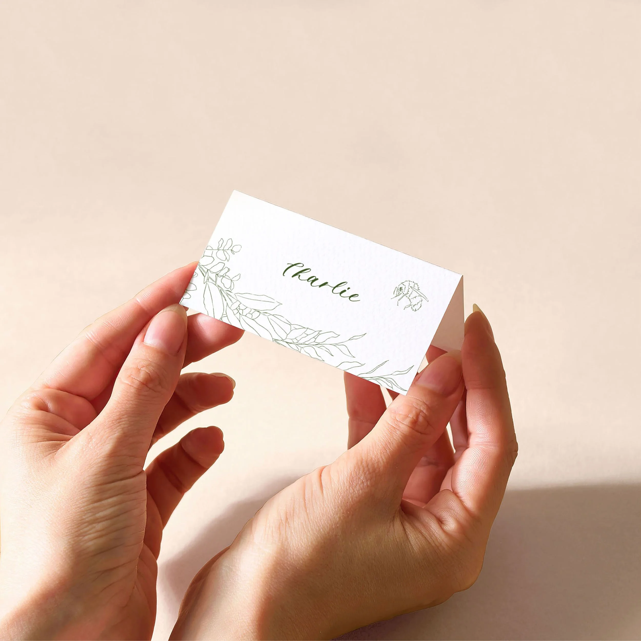 Hands holding a white tent fold place card with a guest name printed on it and delicate green leaf illustrations, featuring a small bee.