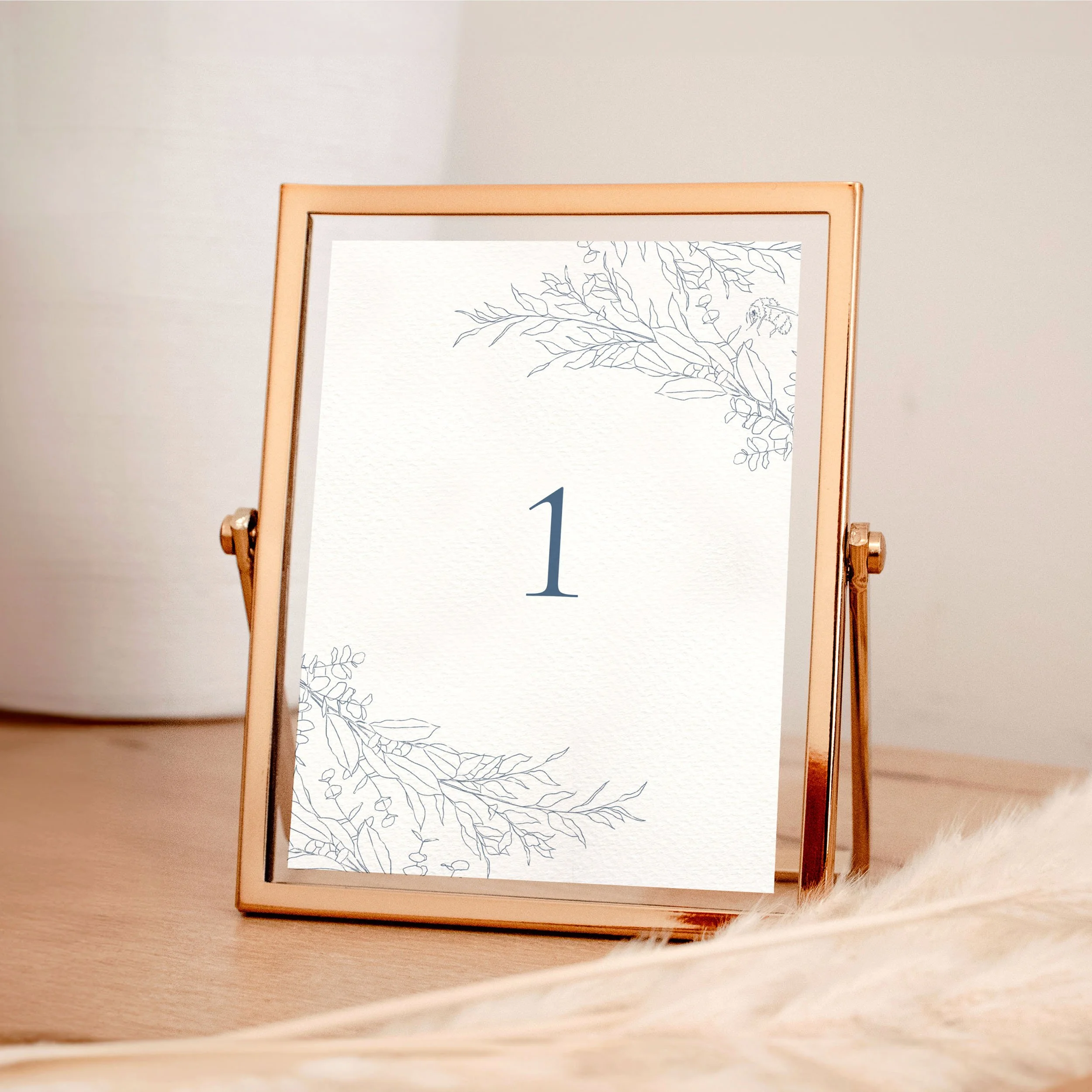 A table wedding number card in a rose gold frame with dusky blue hand-drawn leaves framing the top and bottom