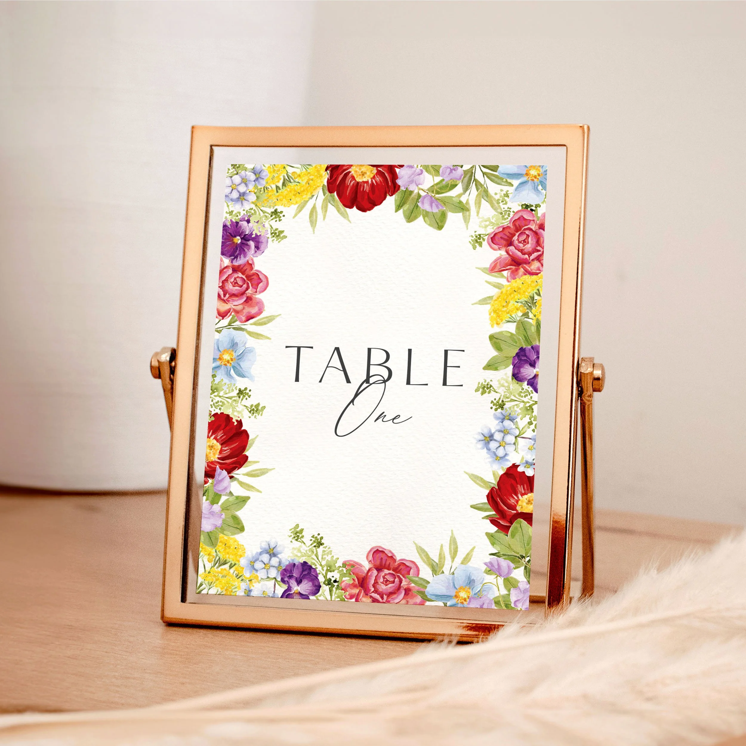 A table wedding number card in a rose gold frame with bright colourful illustrated flowers framing the edges