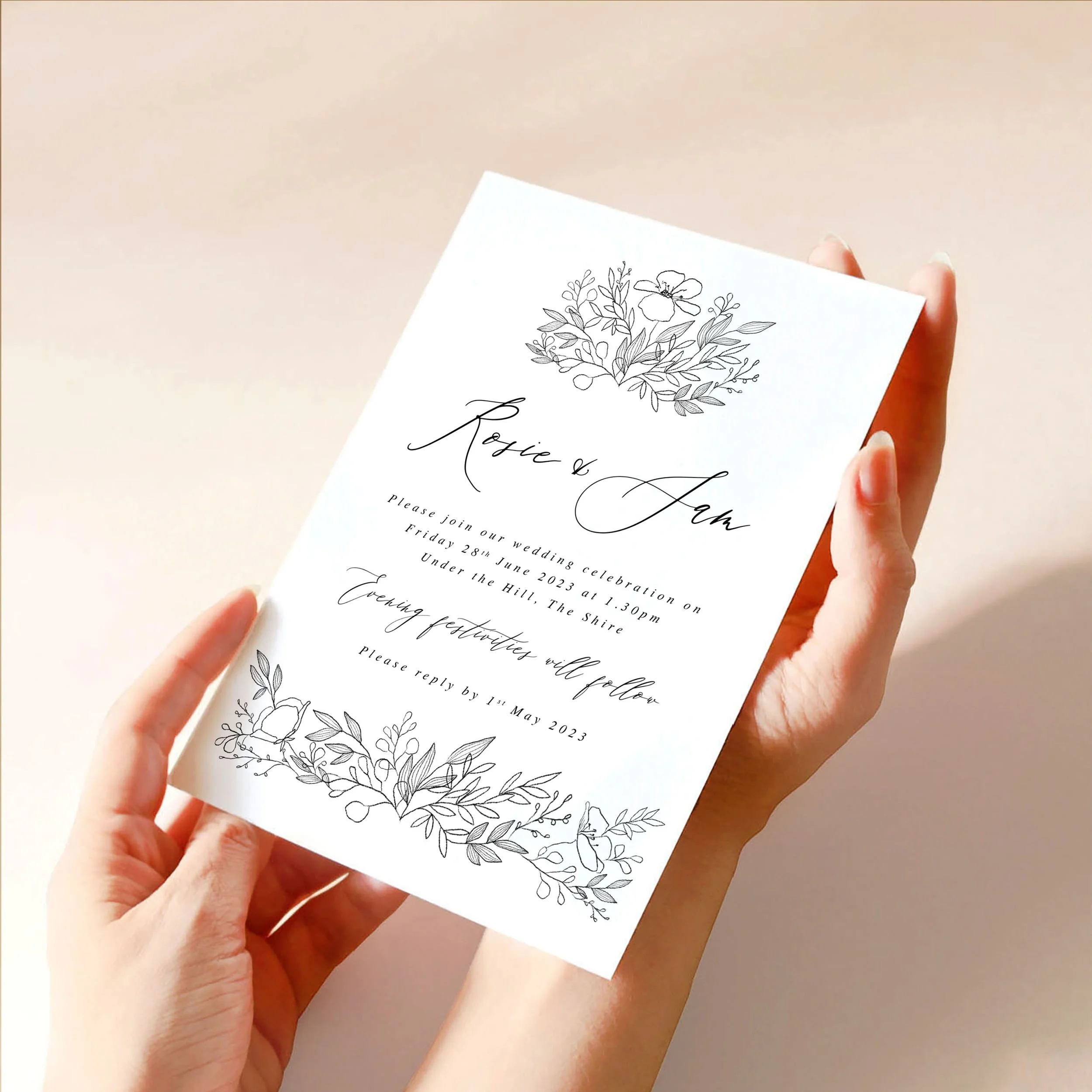 Two hands hold a 5x7" wedding invitation card. It has a hand-drawn sketchy motif at the top and bottom in black and calligraphy-style text.