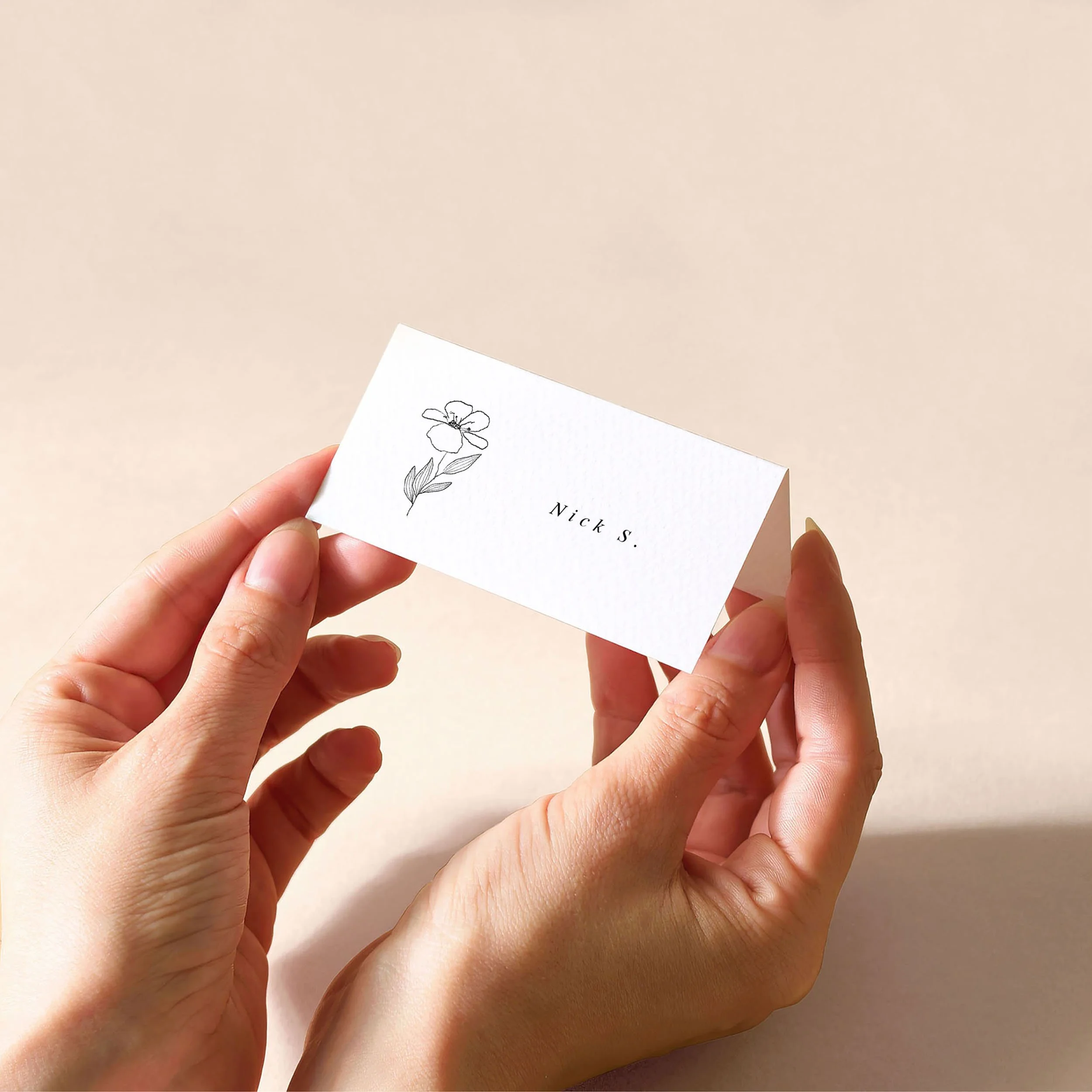 Hands holding a white tent fold place card with a guest name printed on it and a small illustrated black flower.
