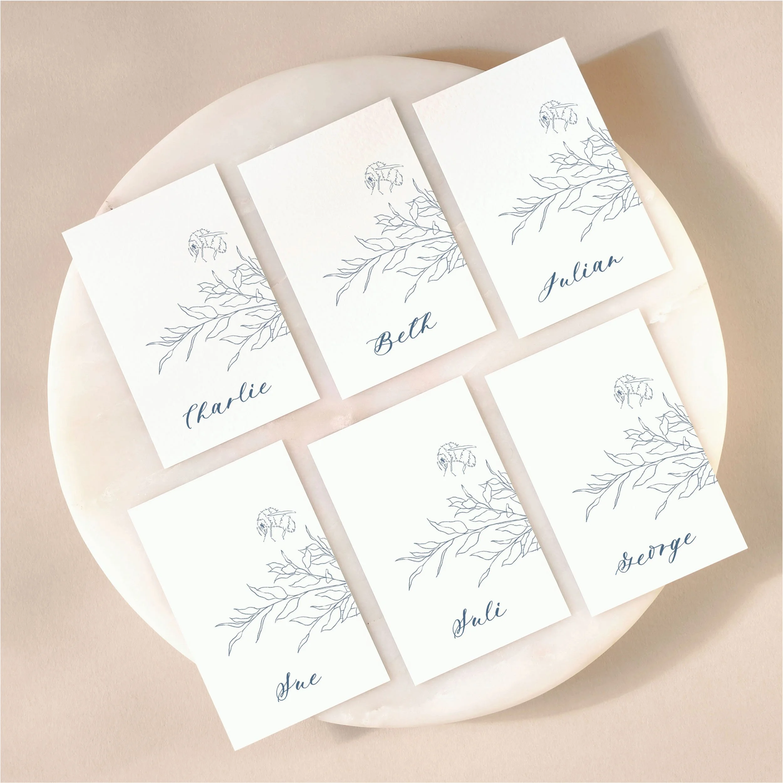 A set of 6 small, flat wedding place cards on a marble tile with printed guest names featuring an illustration of some leaves and a bumble bee