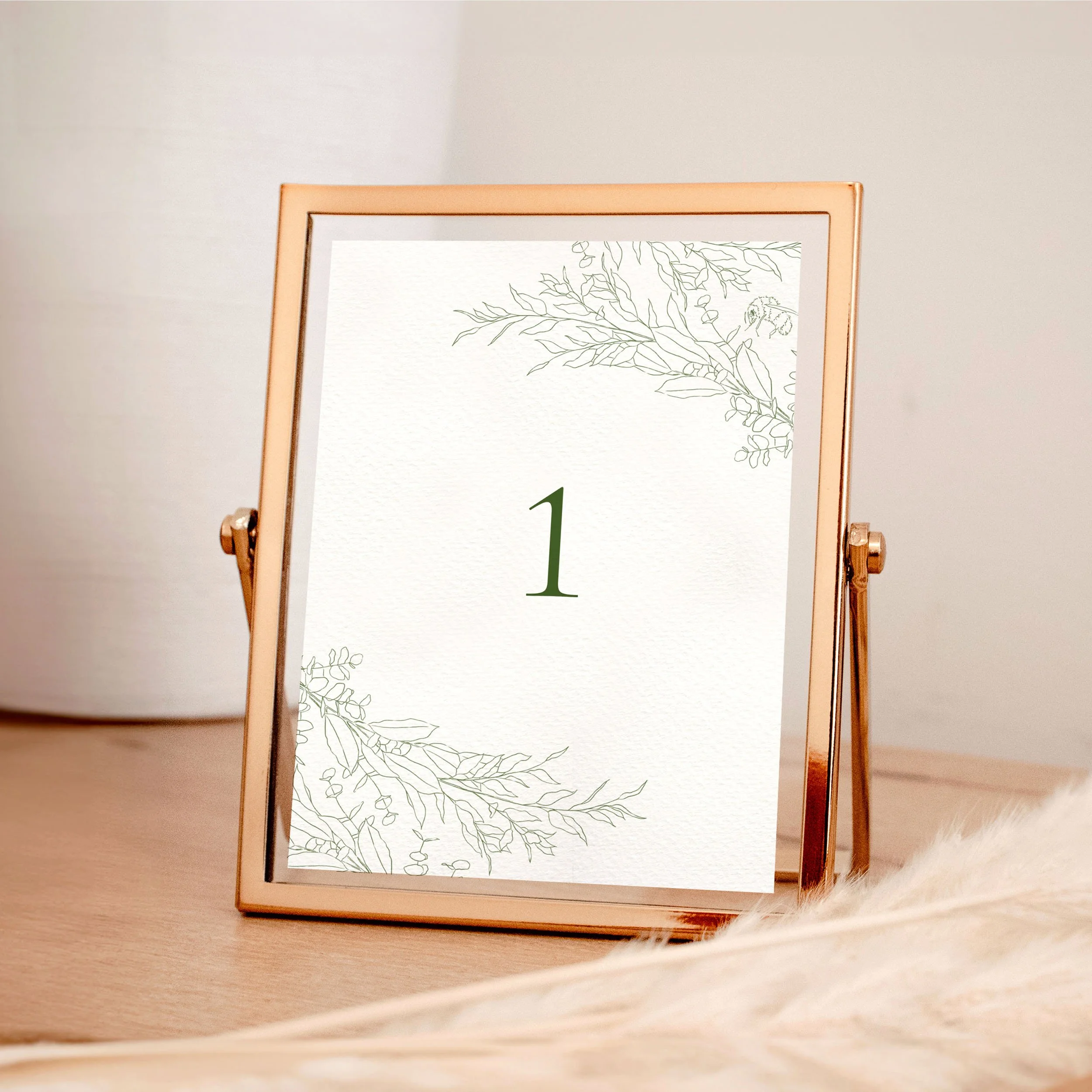 A table wedding number card in a rose gold frame with green hand-drawn leaves framing the top and bottom