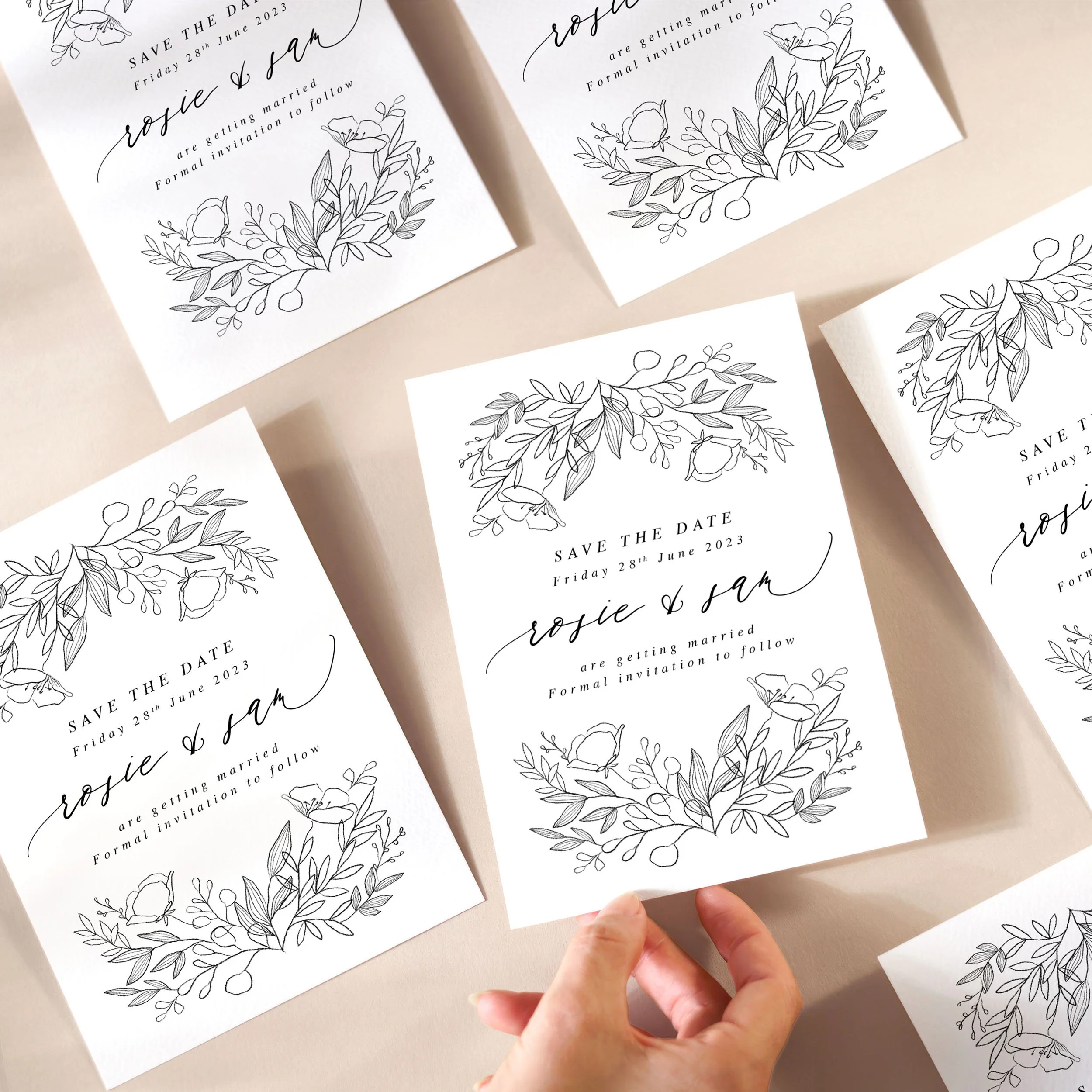 A hand picking up a save the date card with a monochrome floral sketch at the top and bottom, and a modern mix of serif and calligraphy-style fonts.