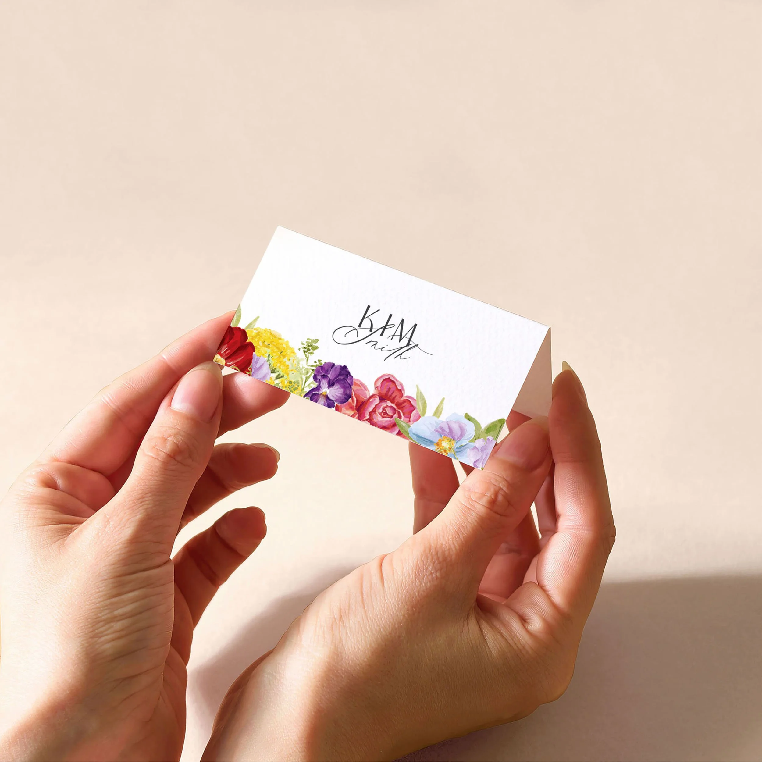 Hands holding a white tent fold place card with a guest name printed on it and a bright hand-painted flowers.