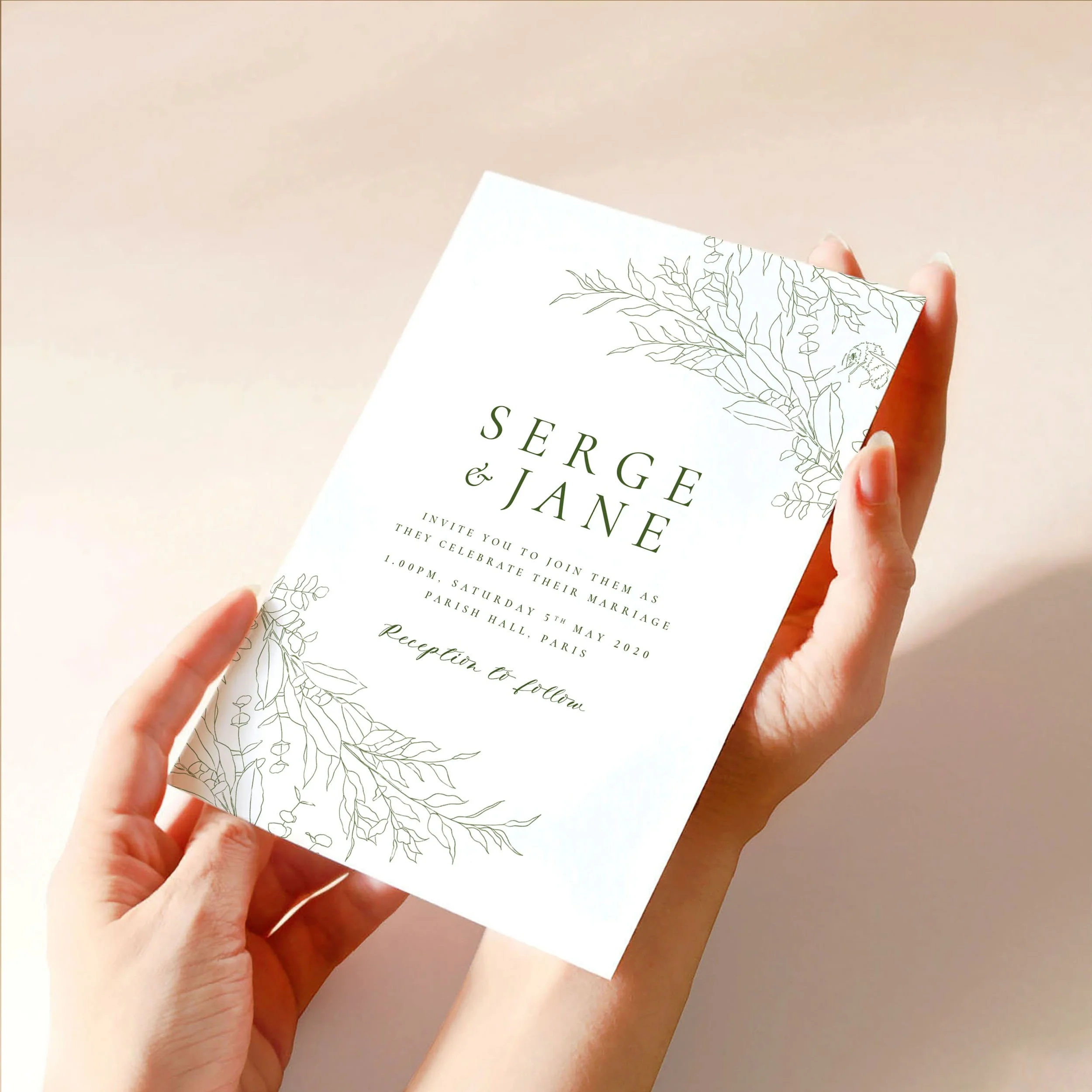 Two hands hold a 5x7" wedding invitation card. It has a green hand-drawn leaf design with a bumble bee at the top and bottom and calligraphy-style text.