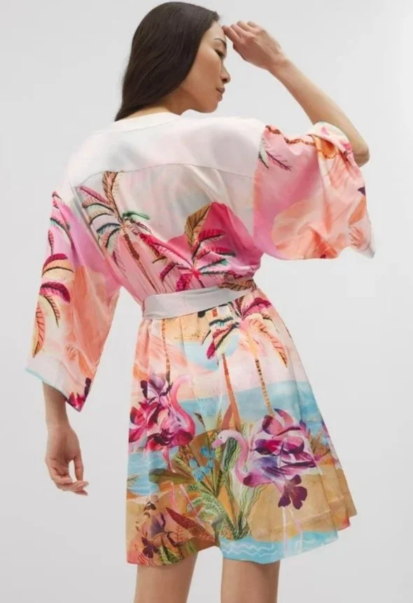 Woman wearing a colorful tropical-themed dress with flamingos and palm trees, standing with her back to the camera, in a white studio background.
