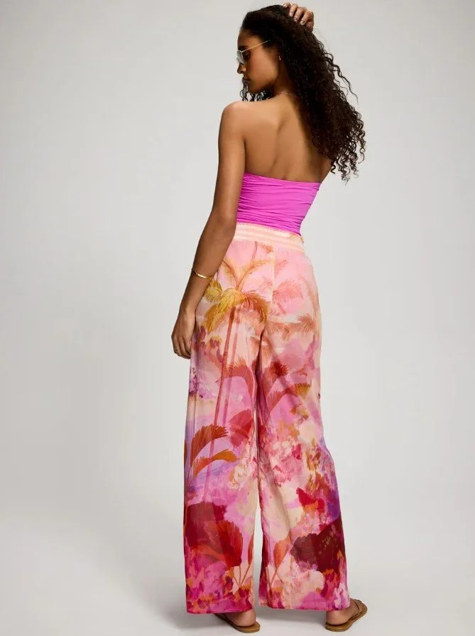 A woman with curly hair wearing sunglasses, a pink strapless top, colorful wide-leg pants with a pink, orange, and red tropical pattern