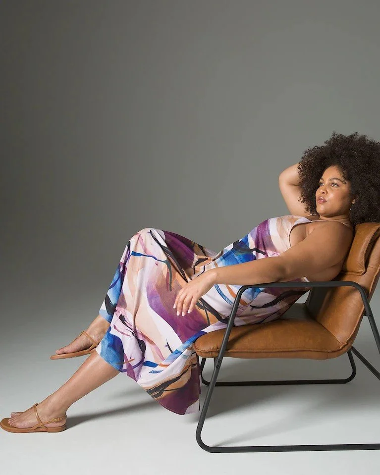 A woman with curly hair lounging on a modern brown leather chair, wearing a colorful patterned dress, with one arm resting behind her head and the other on her lap, against a plain gray background.