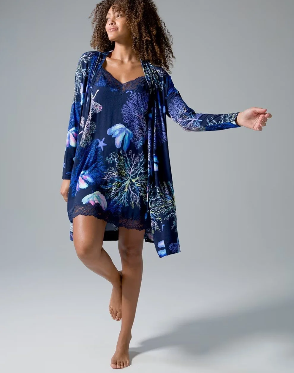 Woman with curly hair in a blue ocean-themed dress and robe, standing barefoot against a gray background.