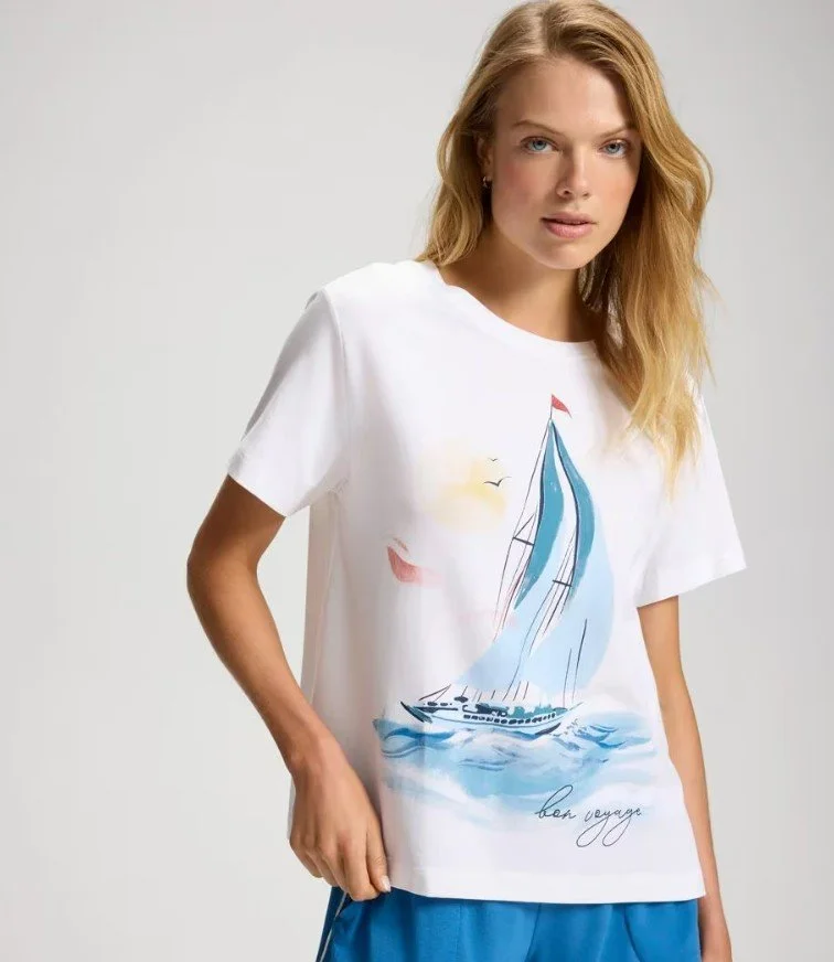 A woman wearing a white t-shirt with a colorful sailboat graphic and the words 'bon voyage' written underneath, standing against a plain light background.